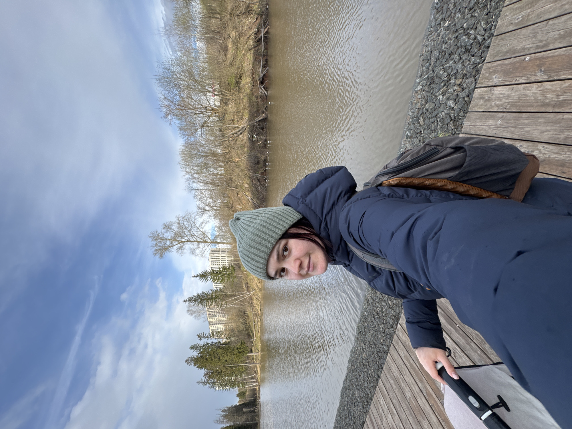 This sideways portrait image captures a young woman smiling faintly and looking directly at the camera. She wears a light green knit beanie, a dark blue puffer jacket, and a dark-colored backpack on her shoulders. Her right hand is visible holding the handle of what appears to be a baby stroller or carriage. She is positioned on a wooden boardwalk, which runs alongside a strip of grey gravel.
Behind her is a wide body of murky, brown-green water, indicative of a river or lake. The opposite bank is lined with a mix of bare deciduous trees and some evergreen conifers. Several white birds are visible on the water's surface near the far shore. In the distance, behind the trees, multiple light-colored multi-story buildings are visible, suggesting an urban or suburban environment. The sky above is partly cloudy with patches of blue. The bare trees indicate an early spring or late autumn season. The overall scene depicts a calm outdoor setting, likely a park or promenade by the water in Izhevsk, Russia. The woman's expression suggests contentment. No text is visible in the media file.