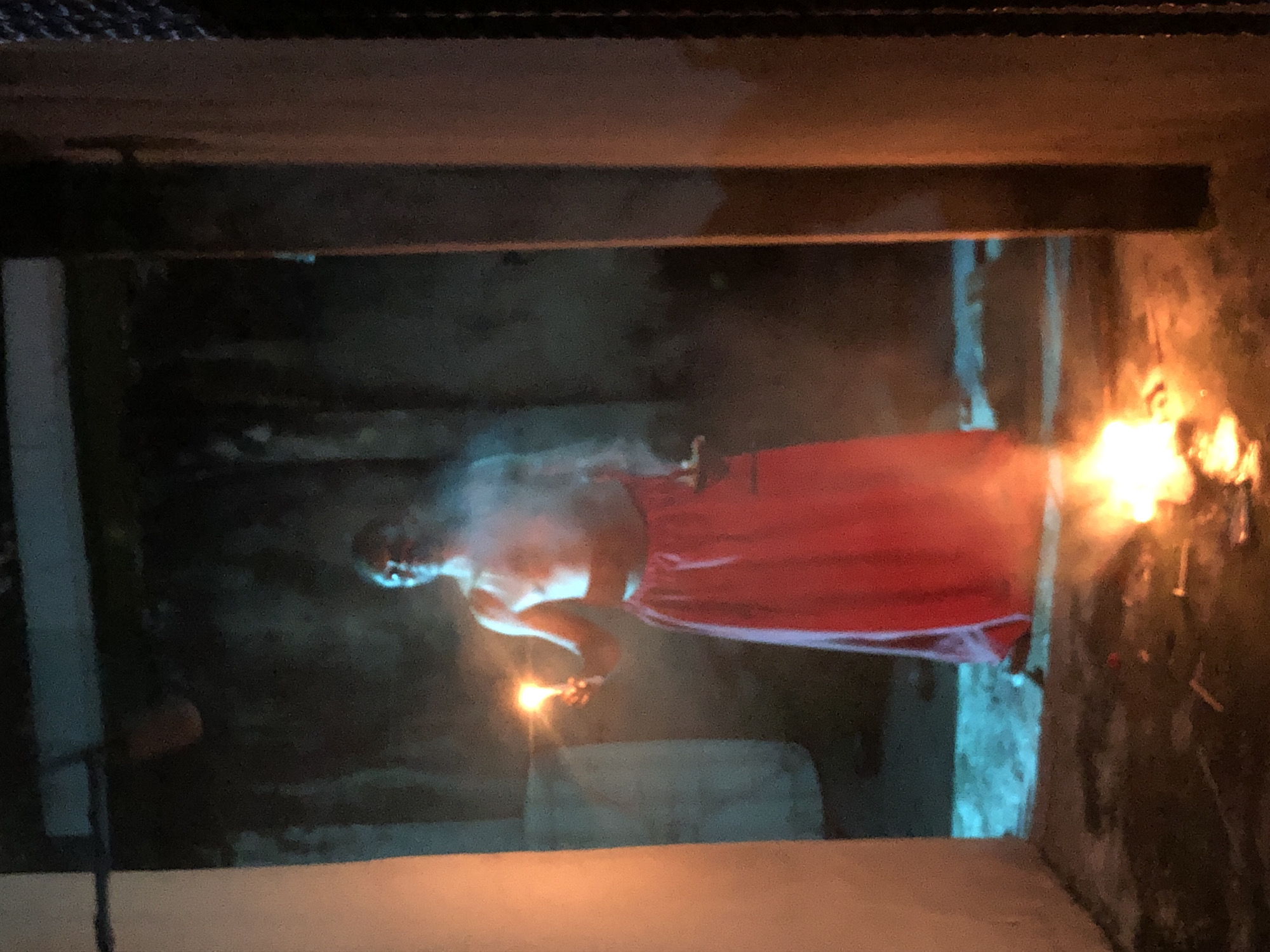 A person, wearing a red wrap-like garment, stands within a dark, box-like structure. The person holds a small torch or light source in their left hand. A larger fire burns on the right-hand side, illuminating part of the structure. The environment appears to be enclosed with concrete-like walls or supports.