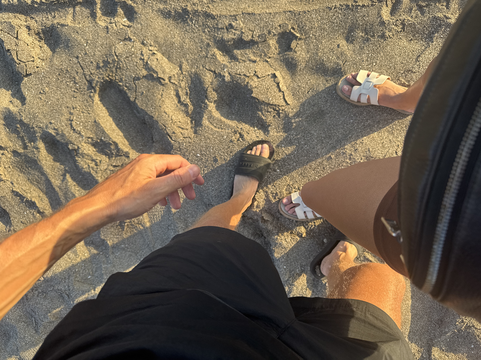 The image, seemingly taken from a first-person perspective, features two individuals standing on a sandy surface, likely a beach. The location is North Kuta, Indonesia, identified by the presence of dark volcanic sand common in the region.
The person taking the picture is wearing black shorts and dark-colored slides. Their left arm and hand are visible in the frame. Another person, likely a woman, is wearing brown shorts and white sandal shoes. A dark bag is positioned near their right side.
The sand shows visible footprints and is illuminated by strong sunlight, casting shadows across the frame. The overall impression is that the individuals are casually standing on the beach.
