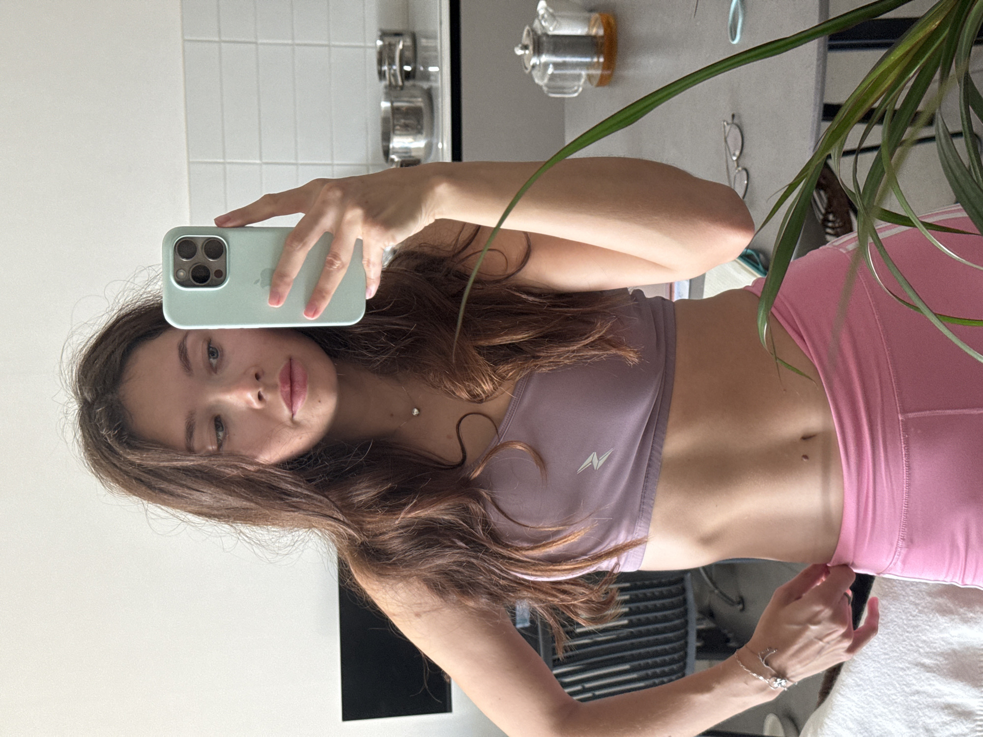 A young woman is captured in a mirror selfie, positioned vertically in the frame. She holds a light green-cased iPhone, likely a Pro model, with her right hand, extending her arm to take the photo. Her long, dark brown hair flows down her shoulders and across her chest. She wears a lavender sports bra featuring a white, stylized 'N' logo on the left side, paired with pink high-waisted athletic leggings or shorts. A small, silver necklace with a pendant and a silver bracelet with charms (possibly a moon and star) adorn her. Her expression is neutral with slightly parted lips, and her light-colored eyes look towards the camera's reflection.
The background, visible through the mirror, suggests an indoor setting, possibly a kitchen or an adjacent area. White tiled walls are visible behind her head. To her left, several silver pots or containers are stacked on a shelf, and further right, a clear glass teapot with a filter is present. Eyeglasses rest on a surface in the mid-ground, and a green houseplant partially obscures the right side of the image. The lighting appears bright and natural, indicating daytime. The word "iPhone" is visible on the back of the phone case.