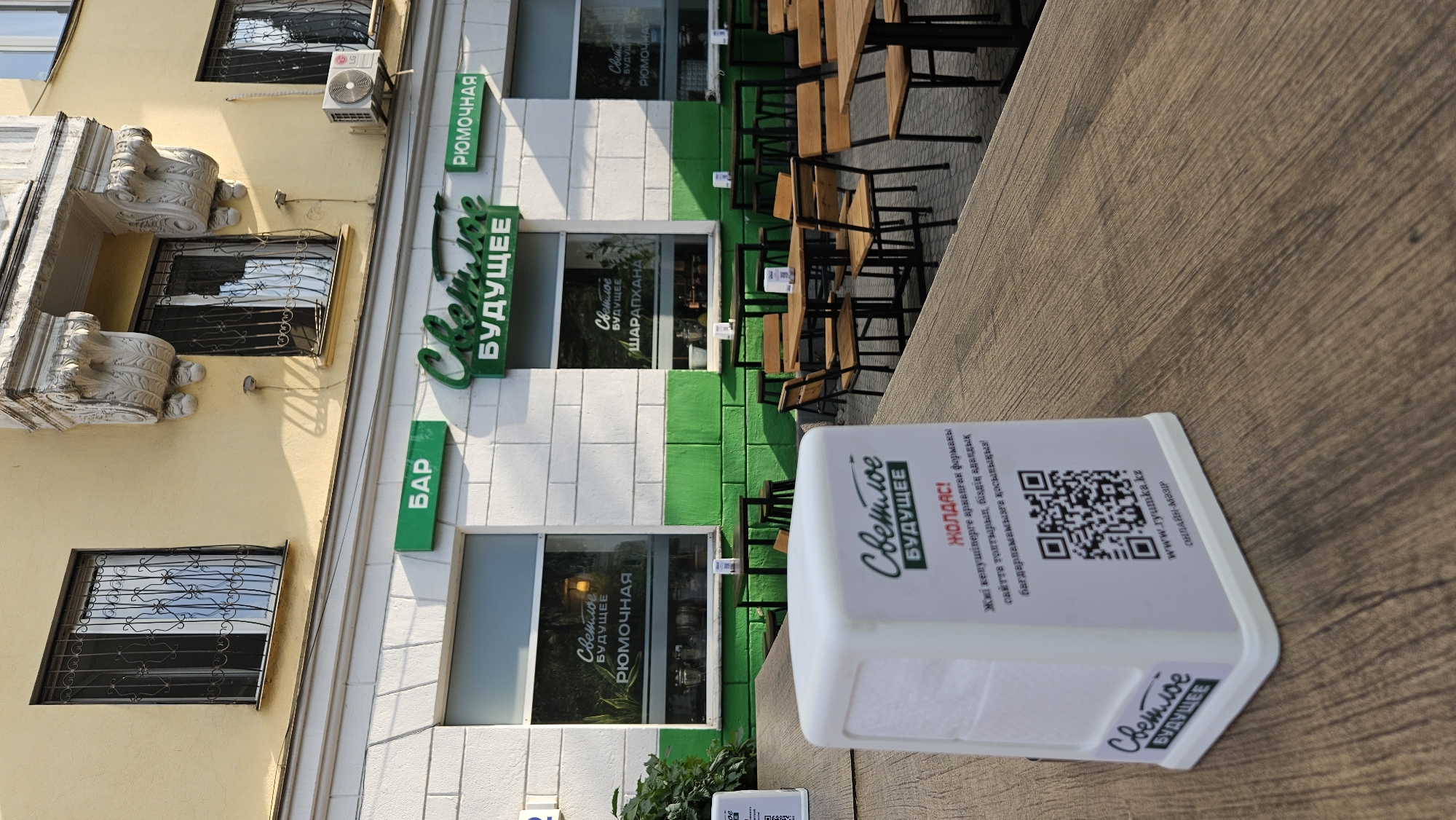 A bright, sunny outdoor scene of a cafe in Almaty, Kazakhstan, features tables and chairs set up on a wooden deck. In the foreground, a white paper napkin dispenser with a QR code and text in Kazakh rests on a table. The dispenser displays the cafe's name, "Светлое Будущее" (Svetloe Budushchee), meaning "Bright Future." Behind the dispenser, a row of tables with chairs extends towards a building. The building has a yellow facade and white and green accents. Green signs above the windows read "БАР" (Bar), "Светлое Будущее," and "Рюмочная" (Rumochnaya, a place that serves shots of alcohol). Windows display the cafe's interior and some bottles on shelves. An air conditioning unit is visible on the facade of the building. The overall impression is a modern, casual dining establishment.