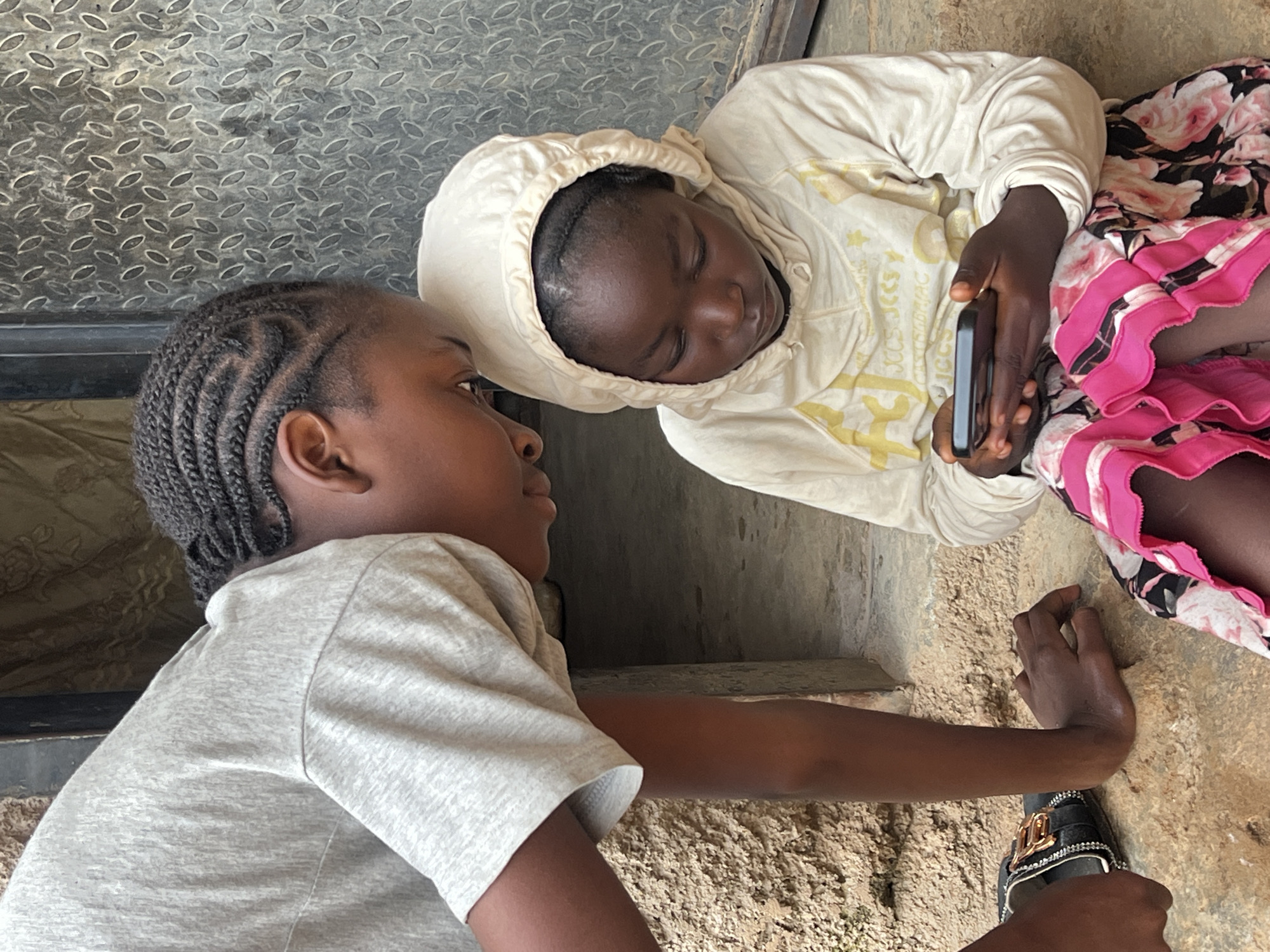 In Jos, Nigeria, two young girls are seen sitting on a rough, textured ground outside a building. The girl on the left, wearing a light gray t-shirt, leans over to look at a smartphone held by the girl on the right. The girl on the right, wearing a cream-colored hooded sweatshirt and a vibrant pink and floral patterned skirt, is focused on the phone. Her hair is styled in neat cornrows, and she is holding the phone horizontally with both hands. The background includes a textured wall and a metallic door with a diamond plate pattern. A sandal with gold embellishments is visible next to the girl on the left. The overall scene depicts a moment of shared attention on a digital device in an outdoor urban setting.