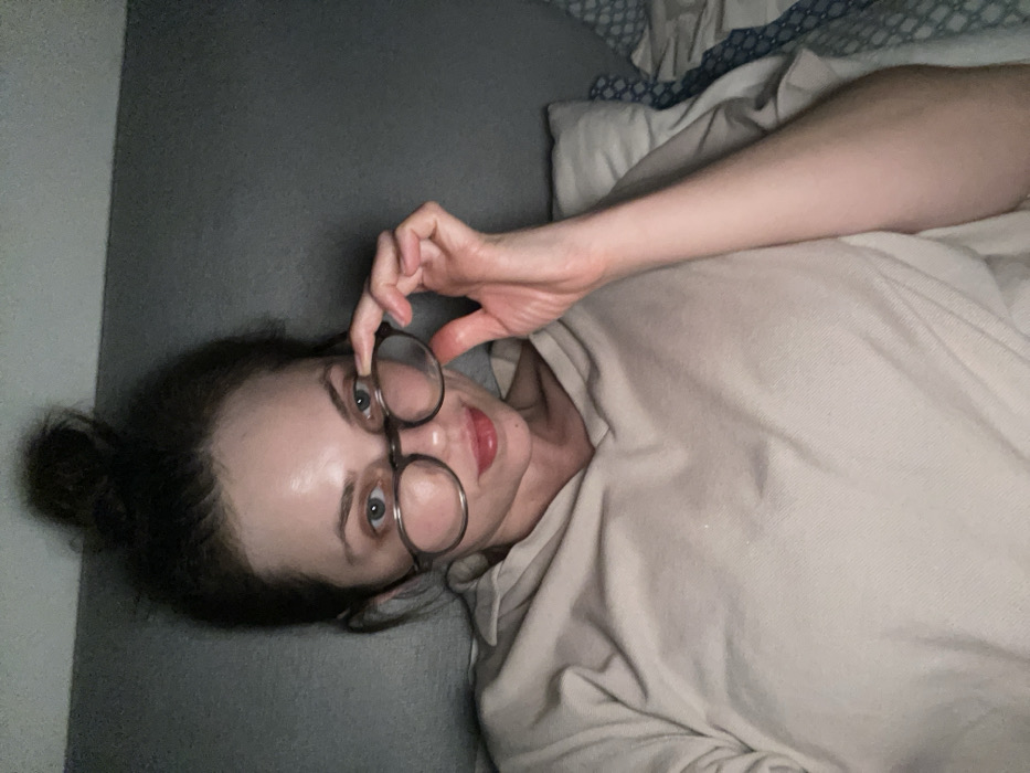 One adult female subject is depicted in a dimly lit indoor setting in Monsummano Terme, Italy. Her dark hair is styled in a high bun. She is wearing a light beige hooded top and round, dark-framed eyeglasses. Her left hand is raised, holding the left temple of the eyeglasses, slightly lowering them from her eyes. Her light-colored eyes are directed at the viewer, and a subtle smirk is visible on her lips, which have a reddish tint. The background consists of a plain dark grey surface, likely a wall or headboard, with a light-colored pillow visible to the subject's right and blue and white patterned bedding visible to her left.