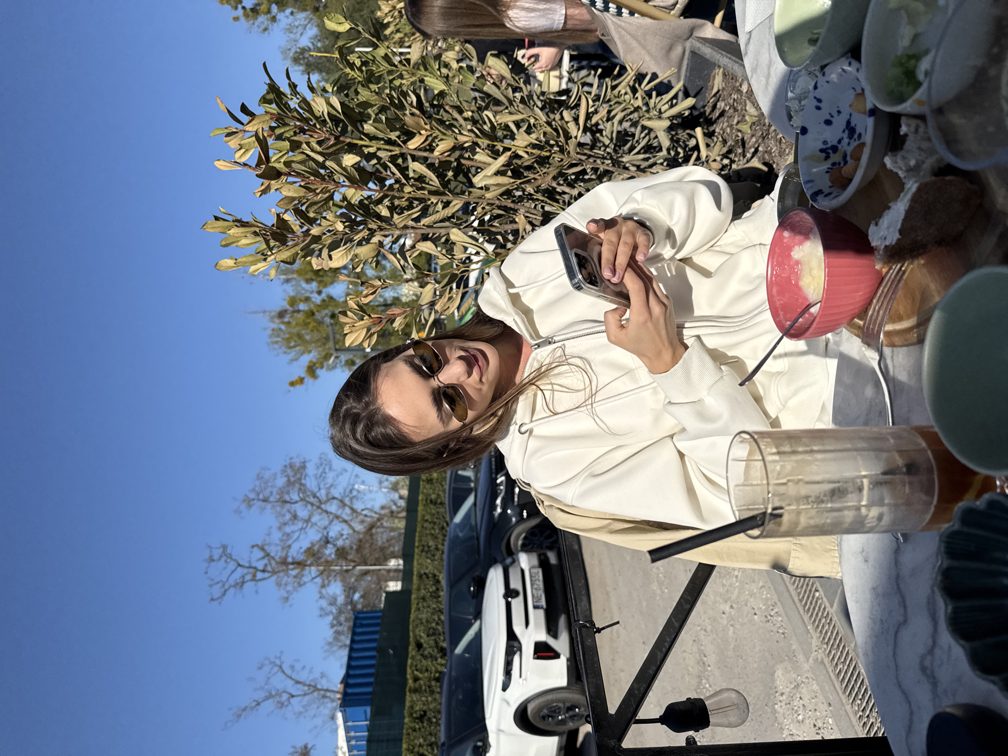 A brightly lit outdoor scene captures a young woman seated at a table, enjoying a meal or snack under a clear blue sky. The image appears to be taken on a sunny day, likely in late autumn or early spring, given the mix of green foliage and bare trees in the background.
The main subject, a woman with long brown hair, is positioned on the right side of the frame. She wears dark sunglasses, a light-colored (possibly cream or white) hooded jacket, and a beige trench coat. Her attention is on a smartphone, which she holds in both hands, encased in a clear cover.
On the table in front of her, a collection of dishes and beverages is visible. A red, ribbed bowl contains a light-colored food item, possibly a type of porridge or yogurt, with a spoon resting inside. Next to it, a tall, clear glass with a dark straw holds an amber-hued drink. Other ceramic bowls and plates, one featuring a distinctive blue and orange speckled pattern, are scattered across the light marble-textured tabletop. A piece of cake or pastry is also present, alongside a fork.
The background features a mix of urban and natural elements. Lush green and brownish bushes separate the dining area from a parking space where several cars are visible. A white SUV clearly displays a Polish license plate, "NE-67554," indicating the location is in Poland, specifically Warsaw as per context. Further back, bare-branched trees stand against the vibrant blue sky, and a blue container-like structure is also discernible. The setting suggests a casual outdoor cafe or restaurant terrace.