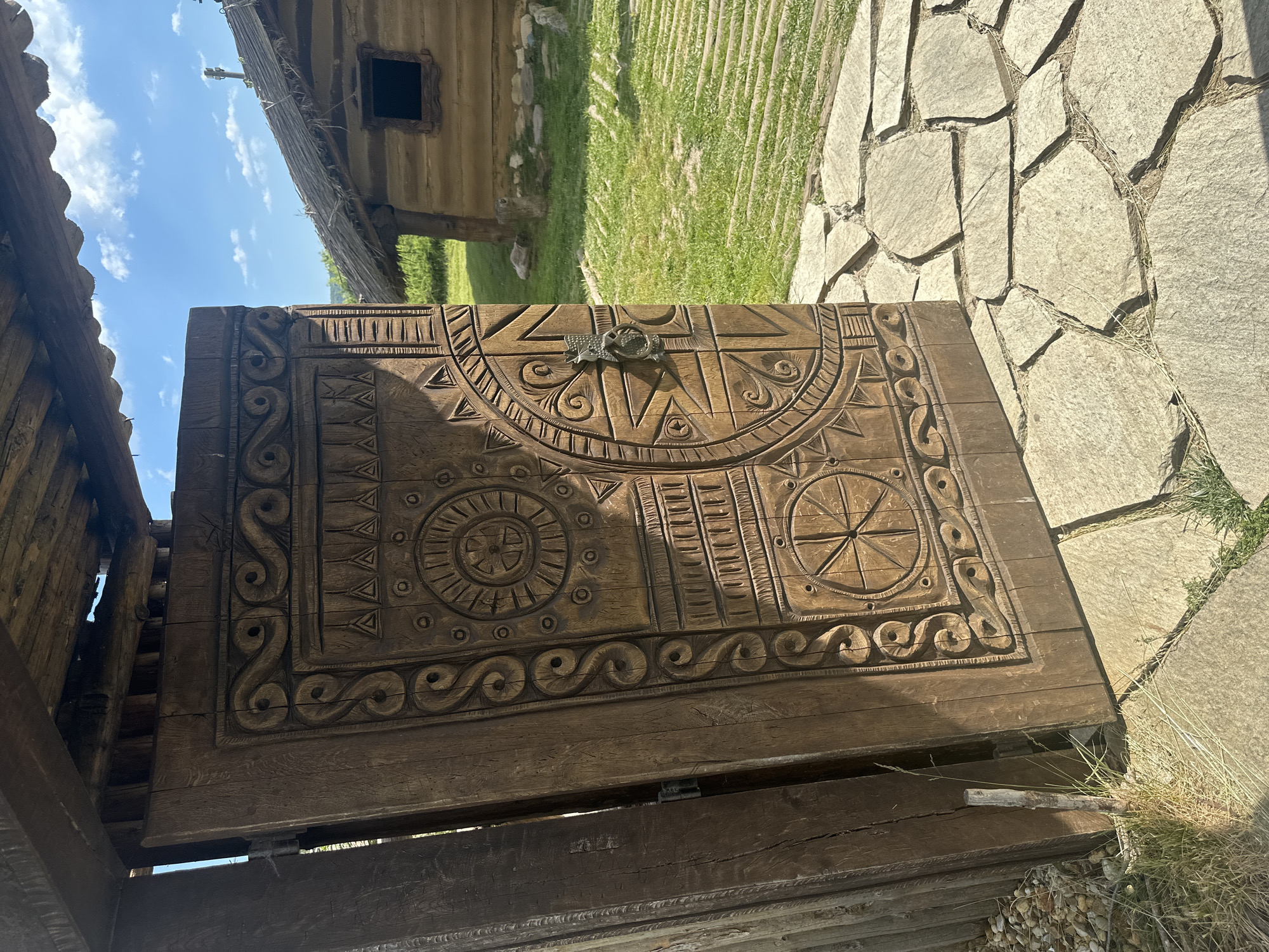 A close-up, eye-level, outdoor shot captures an intricately carved wooden door, weathered and aged, set against a backdrop of a grassy field and a thatched-roof building. The door, adorned with geometric patterns and circular motifs, is partially open, revealing a glimpse of a rustic wooden structure. Sunlight casts shadows across the door, highlighting the detailed carvings. The ground in front of the door is paved with irregular stone slabs, interspersed with patches of green grass and dry straw. In the background, a small, traditional wooden house with a dark window and a chimney stands under a bright blue sky dotted with fluffy white clouds. The scene evokes a historical or traditional atmosphere, possibly within a village or an open-air museum. This image was taken in Neofit Rilski, Bulgaria.