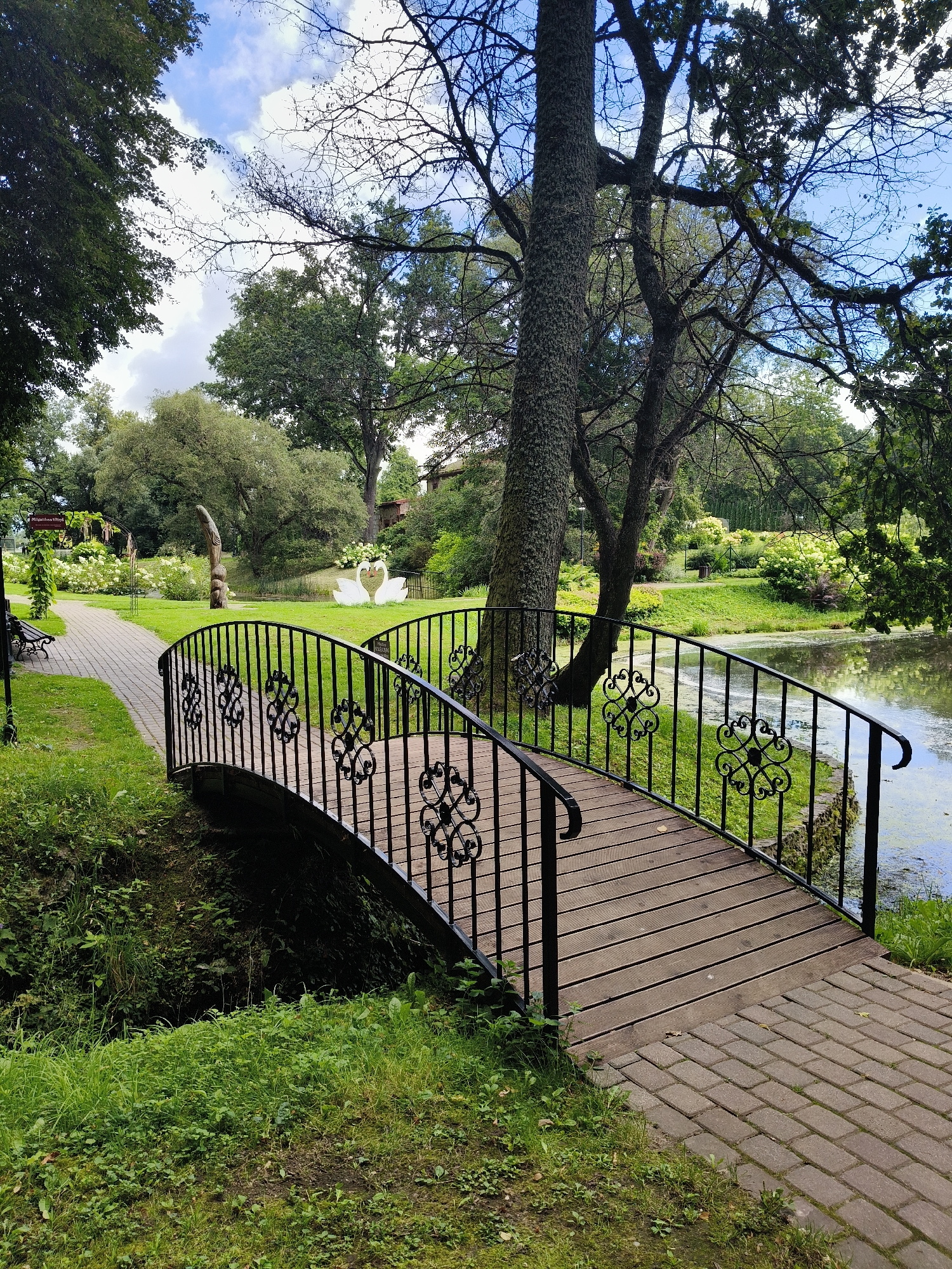 The image shows a park setting in Koknese, Latvia, featuring a small wooden bridge with black wrought-iron railings. The bridge crosses a narrow water channel, with lush green grass and vegetation on either side. Beyond the bridge, a paved walkway leads through the park, past benches and ornamental flower beds. In the background, a pond is visible, adorned with two swan-shaped sculptures. A wooden statue stands near the pond, and mature trees with dense foliage are scattered throughout the landscape. The sky above is partly cloudy with patches of blue.