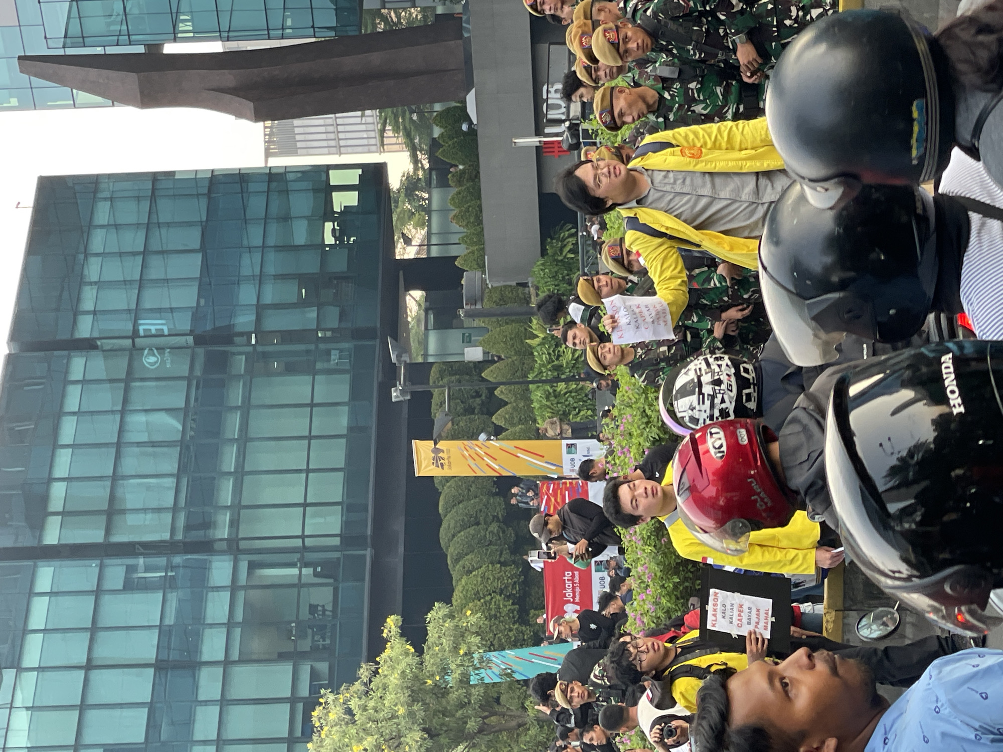 The image depicts an outdoor gathering in Central Jakarta, Indonesia, likely a protest or demonstration. A group of individuals, some wearing military-style camouflage uniforms and berets, are visible in the foreground and midground. In the center, a man in a yellow jacket holds a piece of paper with text written on it, seemingly addressing the crowd. Several people are wearing motorcycle helmets, suggesting a large presence of motorcyclists or participants who arrived by motorcycle.
In the background, modern glass-fronted buildings dominate the skyline, indicating an urban environment, possibly a business district. Banners with text and designs are displayed, adding to the atmosphere of a public event. The overall scene is filled with people, creating a sense of a concentrated gathering in a busy city location.