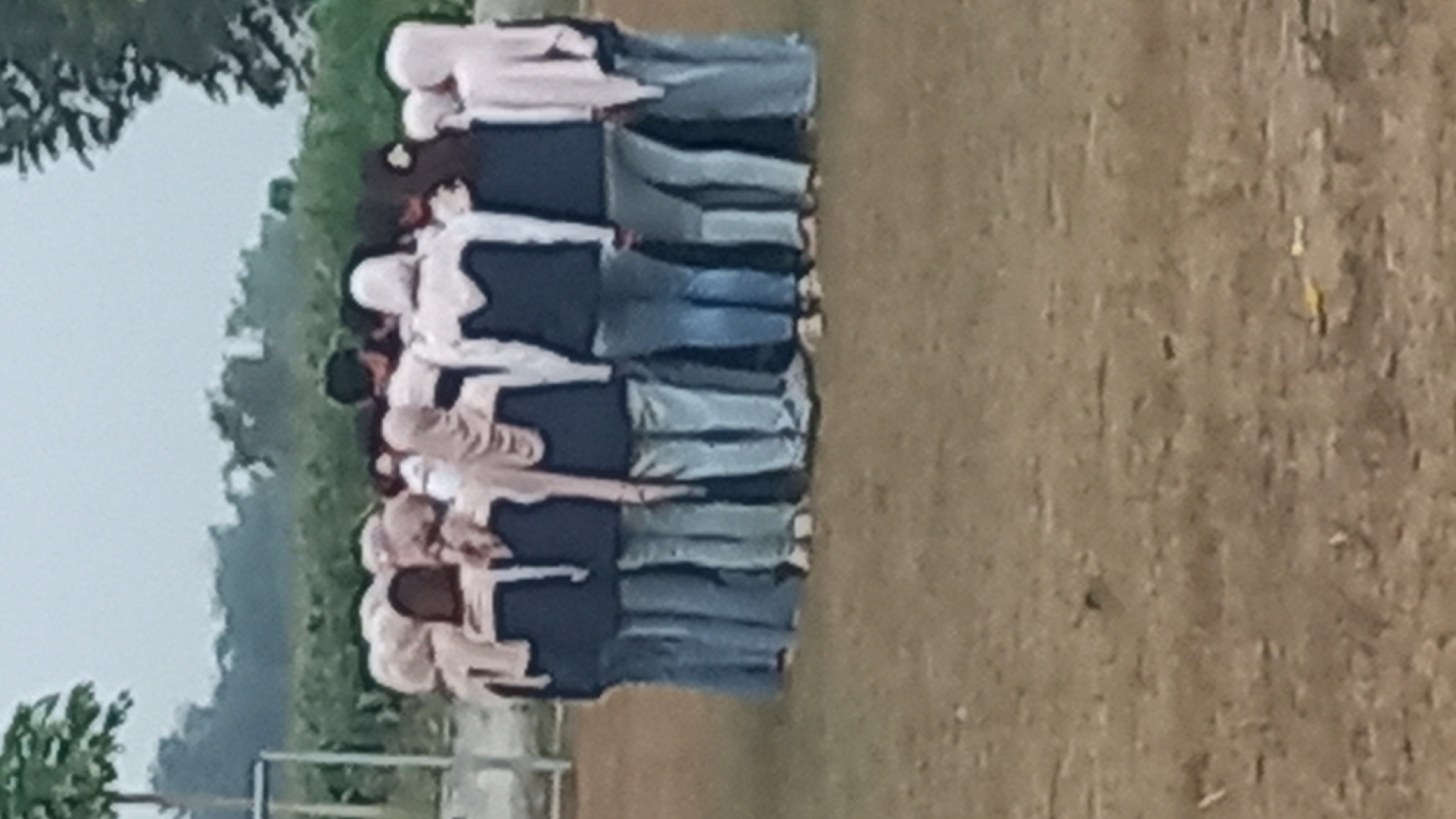 A group of approximately 12 individuals, appearing to be young people, are standing in a circle facing inward on a dirt field. They are dressed in similar uniforms consisting of dark blue vests or jackets worn over light-colored long-sleeved shirts, paired with light blue jeans. Some individuals are wearing headscarves in a similar light color. The background shows greenery, including trees and shrubs, suggesting an outdoor location. The sky is overcast. The image is blurry and low resolution, making precise identification of individuals and details difficult. Based on the known locations in Pare, Indonesia, this scene is consistent with an outdoor school or community activity area.