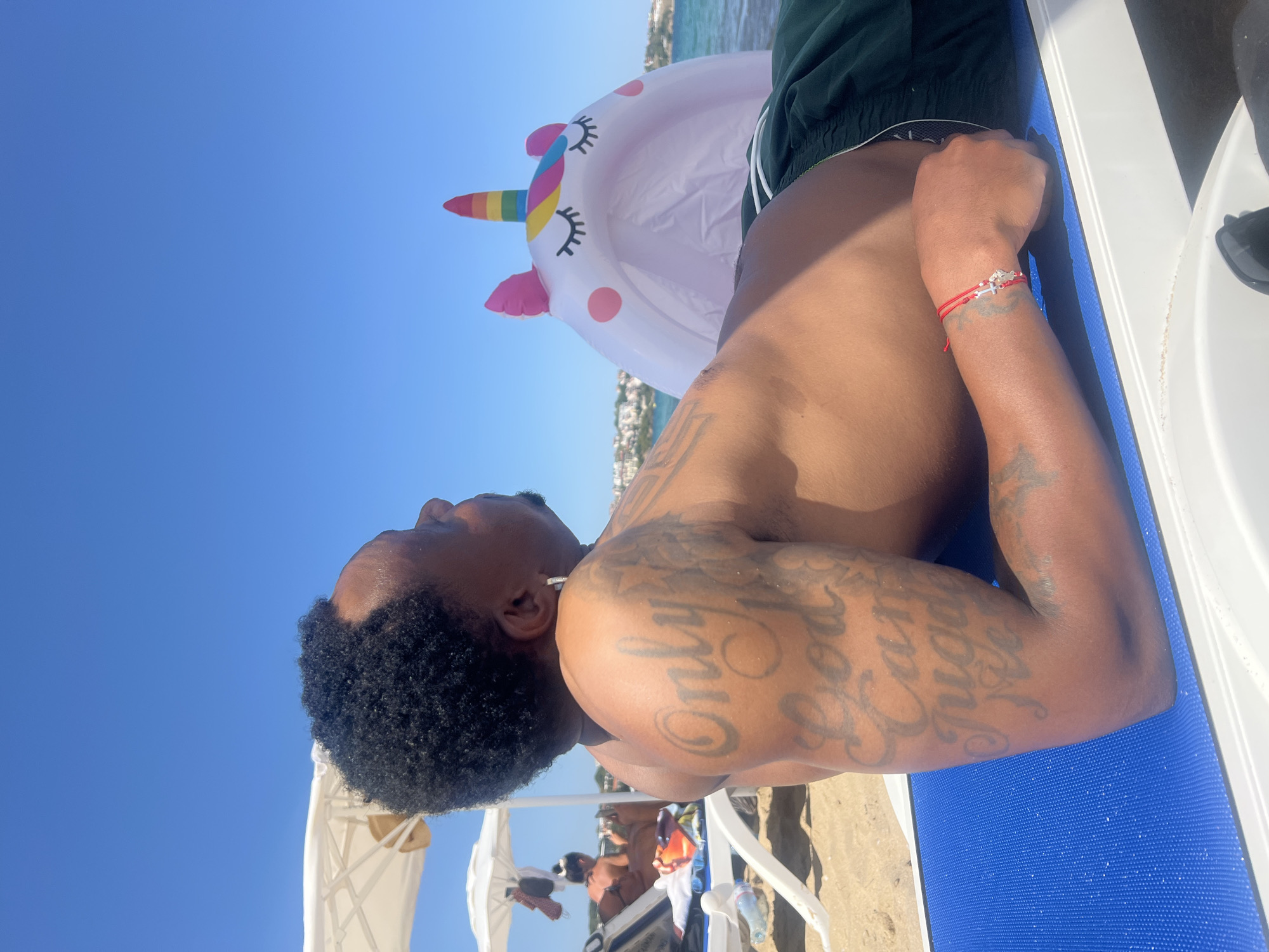 In Sozopol, Bulgaria, a person reclines on a blue lounge chair on a sandy beach. The individual, with dark skin and short, dark, curly hair, is wearing dark green swim trunks. They have numerous tattoos on their left arm, with the words "Only God Can Judge Me" clearly visible. A red string bracelet with a silver charm is worn on their right wrist. Behind them, partially obscuring the view of the sea, is a large, inflatable white unicorn with pink ears and a rainbow horn. The sea is calm, and in the distance, white buildings are visible along the coastline under a clear blue sky. Beach umbrellas and other lounge chairs are present further down the beach.