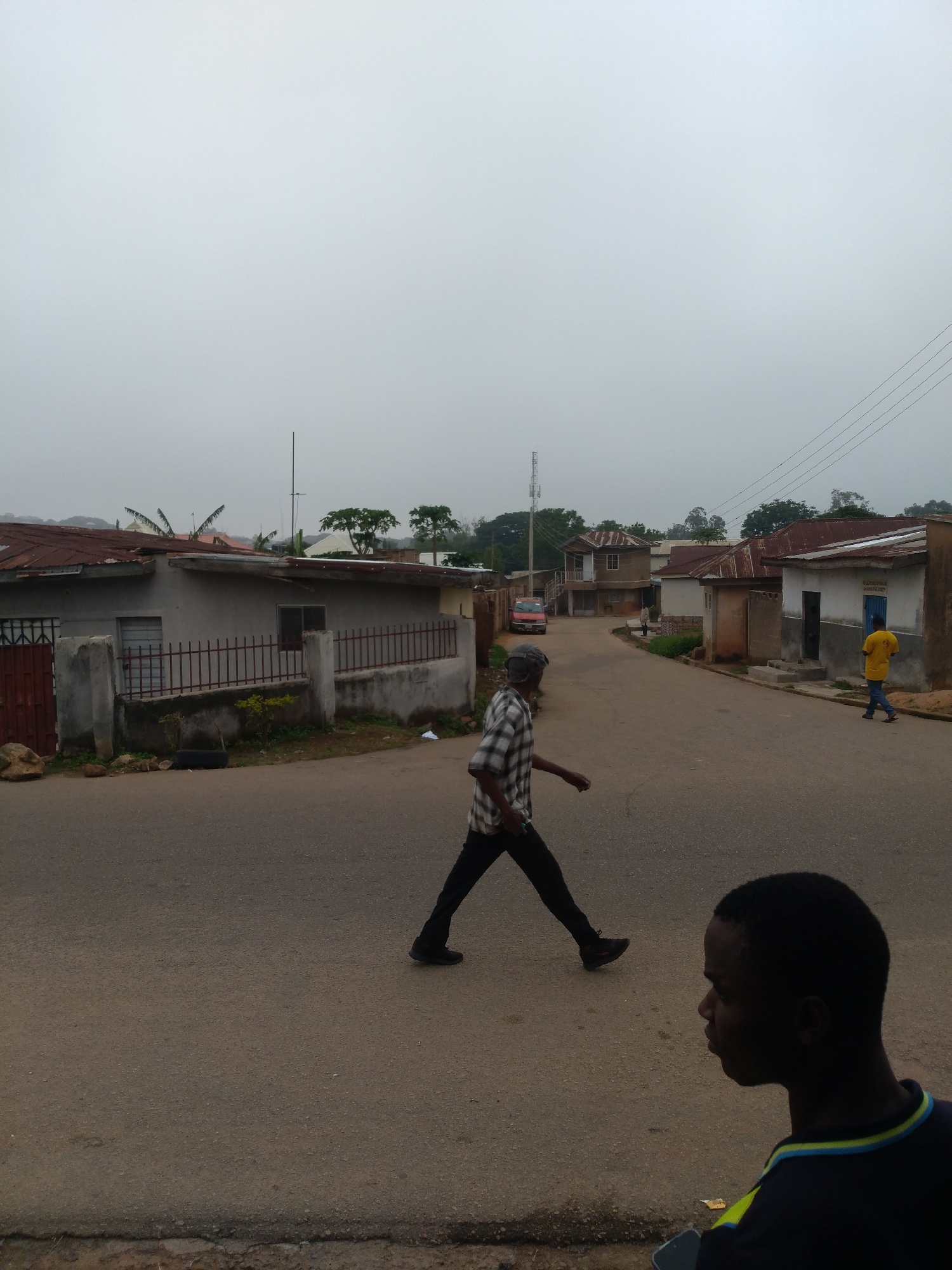 In a street scene of Unknown, Unknown, a man in a plaid shirt and dark pants walks across the asphalt road. Another man, in the foreground and slightly out of focus, looks to his left. Several buildings with metal roofs line the street, which is paved with asphalt and has a concrete curb. Power lines are visible overhead against a cloudy sky. A red van is parked in the distance.
