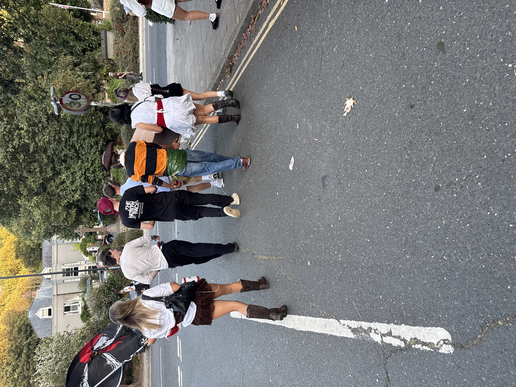 A group of people are walking on a street in St Leonards, United Kingdom. The group includes at least five individuals walking away from the camera. The person in the front left is holding a black and red flag and is wearing a white shirt, brown skirt, and brown boots. To their right, a person in a white shirt and black trousers walks next to another person wearing a black t-shirt and red headscarf. Further back, a person in an orange and black striped shirt and blue jeans carries a brown object. Next to them, a person in a white dress with a red sash walks with a person in a black outfit with a white collar. The people appear to be participating in a parade or event. The environment includes a street with a white line marking, a sidewalk, trees, and buildings in the background. A speed limit sign indicating "30" is visible. The lighting suggests it is late afternoon or early evening.