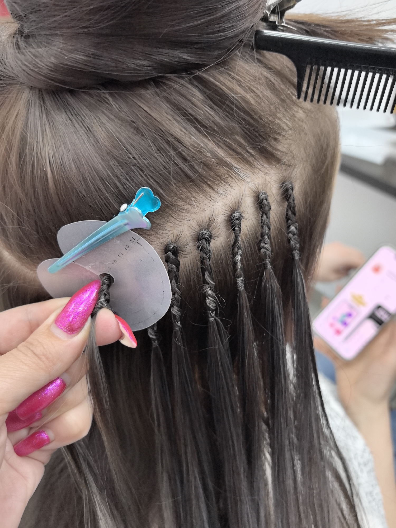 The image presents a close-up view of the back of a person's head during the application of hair extensions. The subject has natural dark brown hair, some of which is sectioned and pulled up with a black comb and clip at the top of the head.
The primary activity is the intricate process of attaching hair extensions. Several small, tightly braided sections of natural hair are visible, each serving as an anchor for the extension strands which hang down. A hand with long, bright pink glitter nail polish is prominent in the foreground, holding a section of hair. This hand is positioned near a white, kidney-shaped protective disc, which features numerical markings "5 10 15 20 25" along its edge. A blue hair clip is used to secure this disc and surrounding hair. The pink-nailed hand appears to be manipulating a new section of hair, preparing it for braiding or extension attachment.
In the blurred background, another person's hand holds a smartphone with a bright pink case, displaying a colorful, likely app-based, interface. The setting appears to be indoors, suggesting a salon or a personal space dedicated to hair styling. The image is a factual depiction of a beauty procedure, without discernible emotions or specific time-of-day indicators.