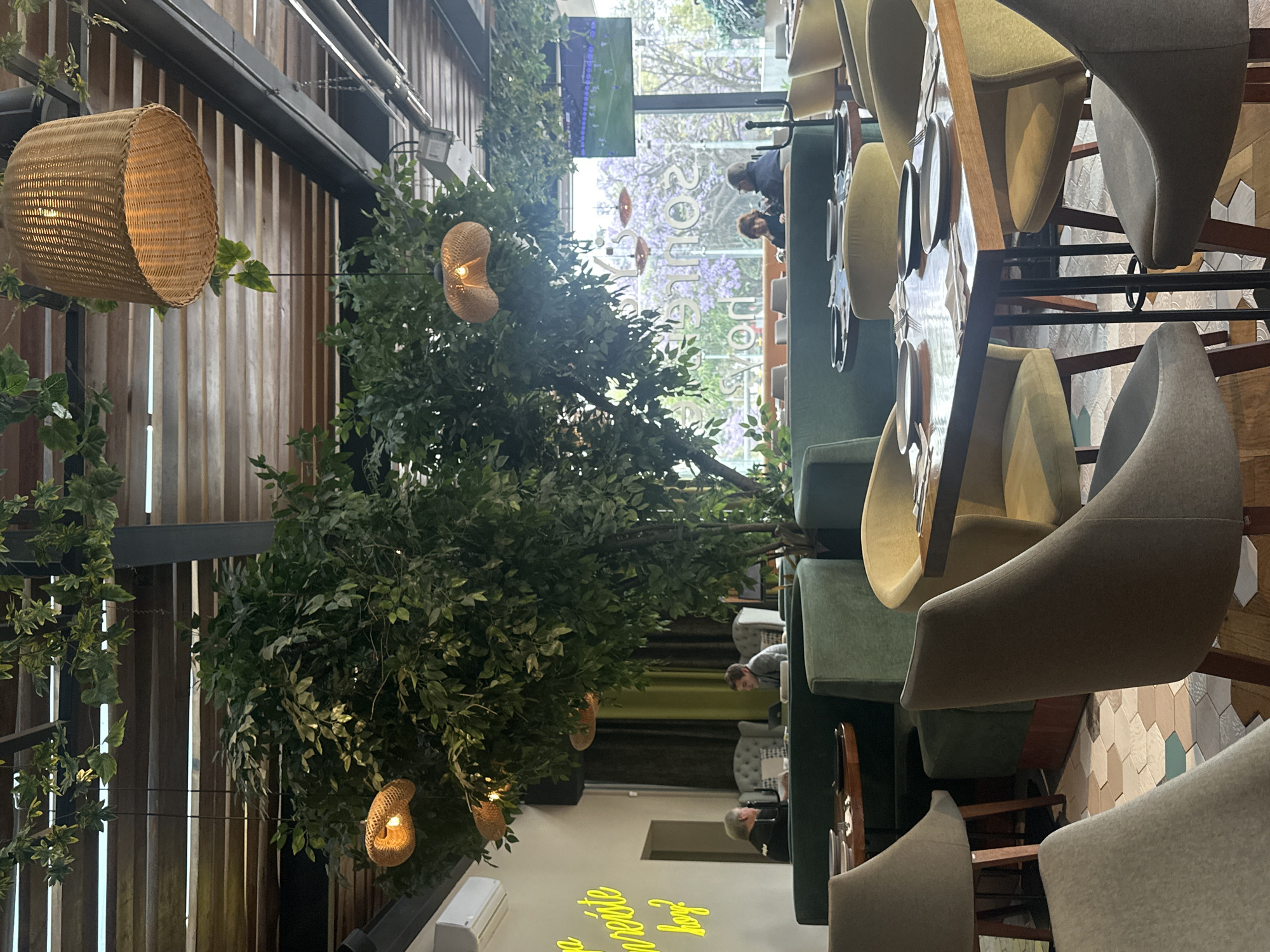 The interior of a restaurant in Huixquilucan de Degollado, Mexico features indoor trees with decorative woven light fixtures suspended from their branches and a metal grid structure. Tables are set along a green booth seating area, with additional modern chairs around the tables. People are seated and appear to be dining in the background. The decor includes wooden walls and hexagon-patterned floor tiles. A neon sign is visible on a nearby wall. Sunlight filters through a large window in the background revealing a view of a tree with purple flowers.