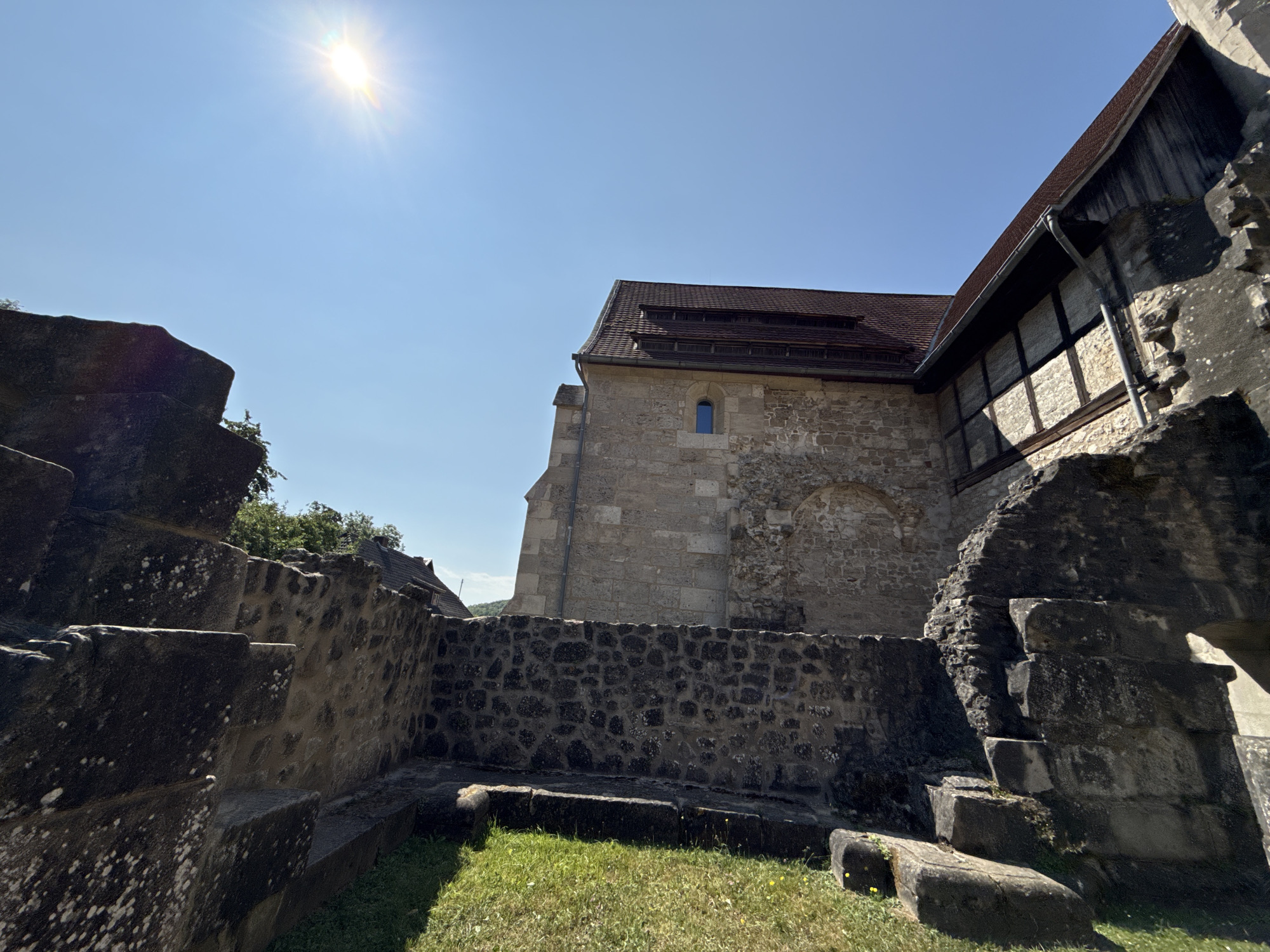 The image shows the exterior of the Walkenried Abbey in Walkenried, Germany, on a clear, sunny day. The foreground features rough, weathered stone structures, possibly remnants of older fortifications or foundations, covered in moss and lichen. These structures are low to the ground, with grassy areas interspersed between them.
In the midground, a well-preserved stone wall made of dark, irregular stones separates the foreground from the abbey building. Behind this wall stands the abbey itself, constructed from light-colored, roughly hewn stones. The abbey features a red tile roof with multiple layers of dormer windows or vents. A small, dark blue-paned window is visible on the stone facade. To the right of the main stone building, another section of the abbey is visible, with a dark wooden facade above the stone, suggesting a timber-framed structure or addition.
The sky is a bright, cloudless blue, with the sun prominently visible in the upper left quadrant, casting a strong glare and lens flare. The sunlight illuminates the scene, creating distinct shadows. In the background, a few trees and the rooftops of other buildings are visible, suggesting the abbey is situated within or near a village or town. No people are present in the image. The overall impression is one of historical architecture set against a bright, natural backdrop.