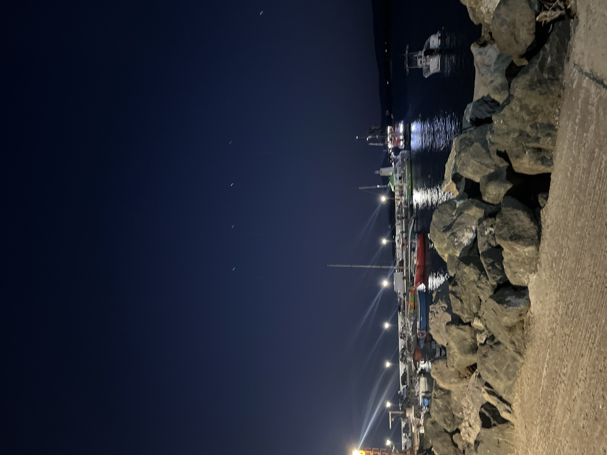 The image depicts a night scene at a harbor, likely in the city of Primorsko, Bulgaria. In the foreground, large, dark rocks line the edge of a paved area. Beyond the rocks, several boats are moored in the water. One boat in the mid-ground appears to be a larger vessel with lights illuminating its upper deck. Other smaller boats, including what looks like a white speed boat, are also visible. The water reflects the lights from the boats and the lights from lamps lining the harbor, creating streaks of illumination. In the background, a dark mass suggests land or hills. The sky above is a deep, dark blue, with several faint, elongated green lights appearing as streaks across it. The overall impression is a tranquil, nocturnal harbor setting.