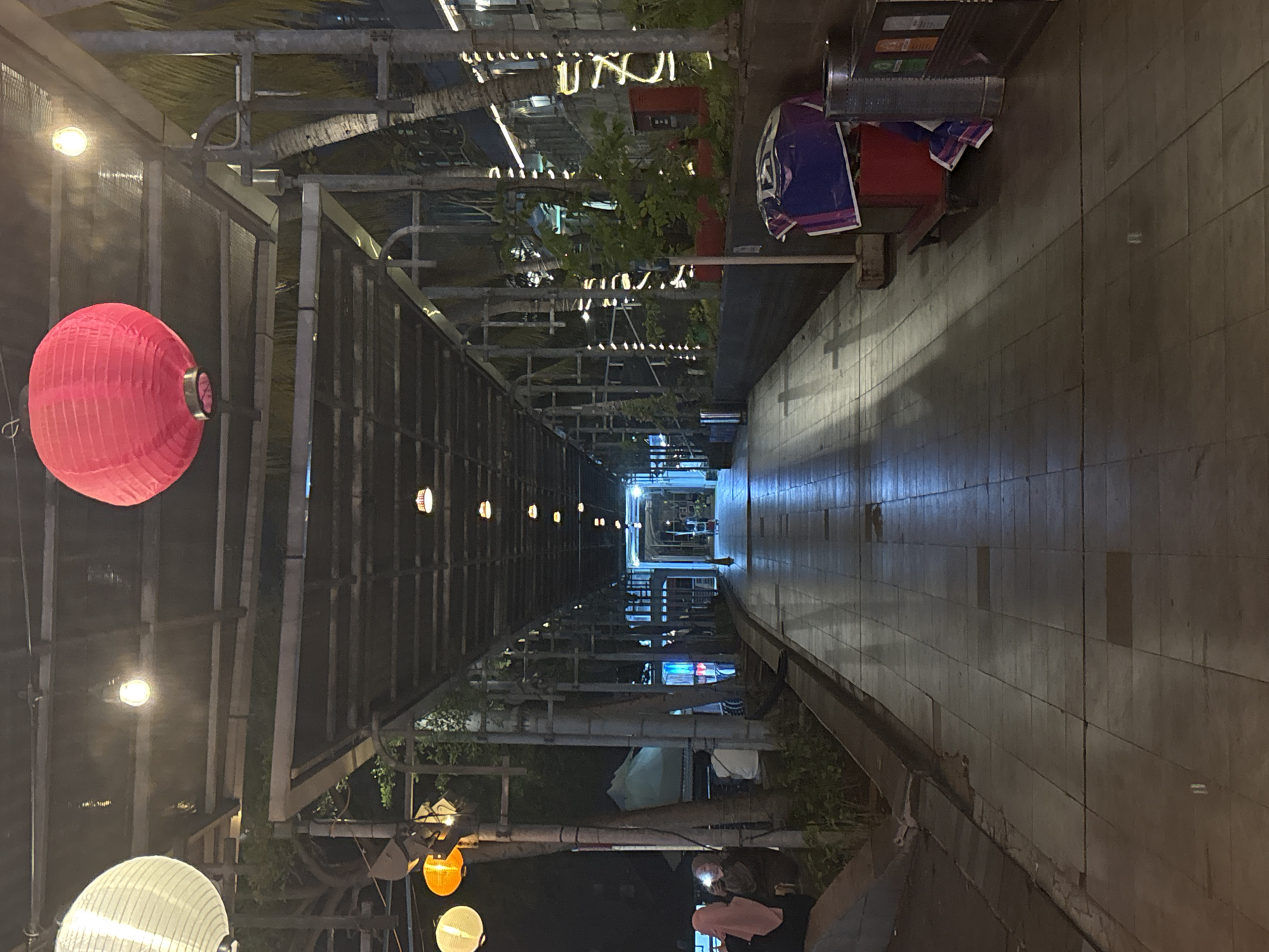 The image depicts an outdoor walkway in North Jakarta, Indonesia, illuminated by a string of lights and traditional lanterns. The walkway is covered by a structure with a dark, slatted roof supported by metal pillars. On the left side of the walkway, several people are seated, with two individuals in the foreground wearing headscarves. Further down the walkway, on the right, are trees with string lights wrapped around their trunks and a red trash receptacle with a blue and white tarp draped over it. The walkway itself is paved with square tiles that appear slightly wet, reflecting the ambient light. In the distance, the walkway opens up to what appears to be a plaza or entrance, also lit by various light fixtures.