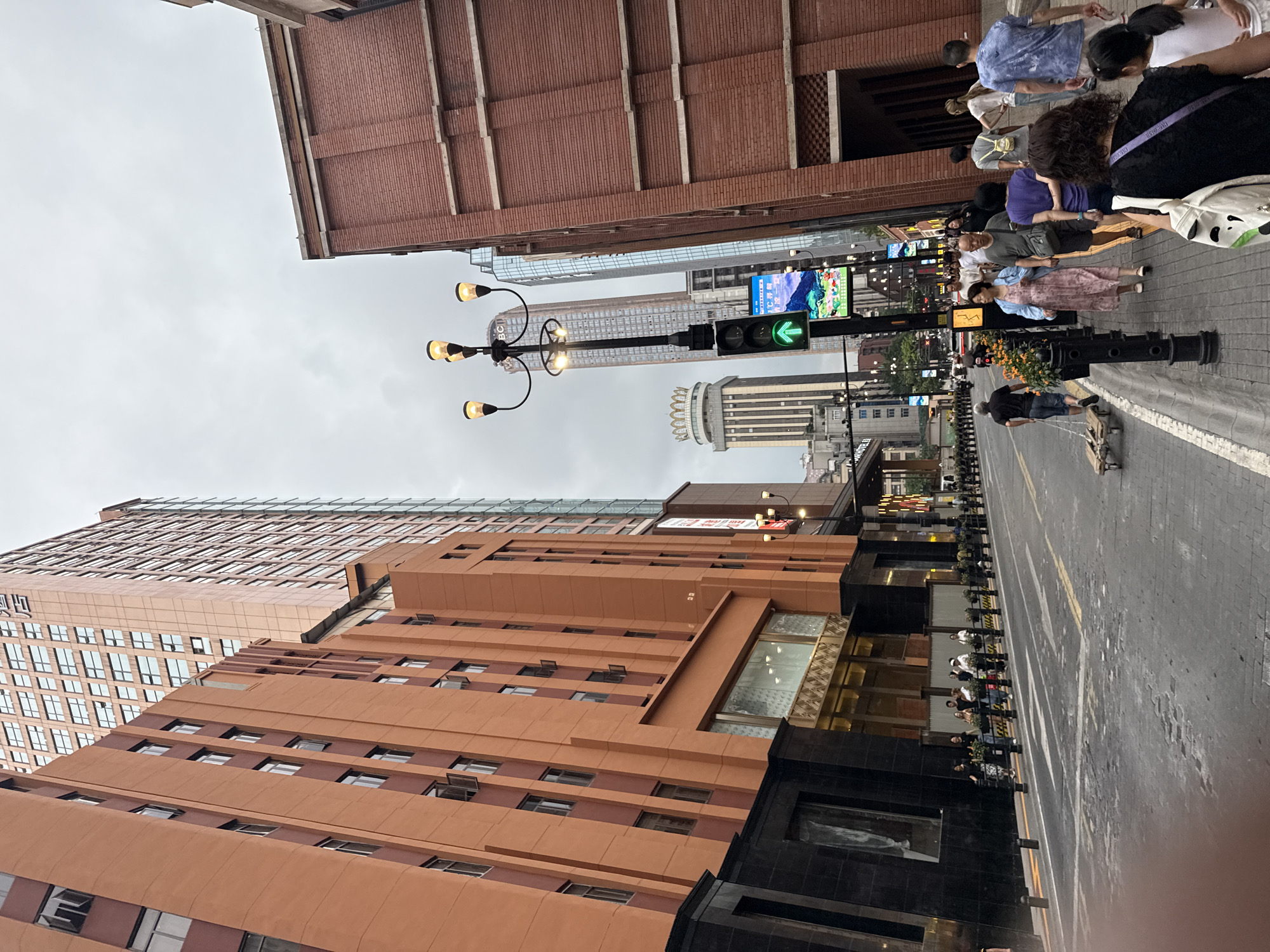 This is a street scene in Yuzhong District, China, on a cloudy day. The camera is positioned at a low angle, looking up towards tall buildings and the sky. On the left, a multi-story building with an orange facade dominates the foreground. It has numerous windows, some of which are open, and a darker stone base. Adjacent to it, another taller building, possibly a hotel with a sign that reads "HOTEL," is visible. Further down the street, a modern skyscraper with a reflective glass exterior stands.
On the right side of the street, a brick building with a reddish-brown facade is prominent. It features horizontal bands of lighter material. Across the street, a series of streetlights with ornate fixtures illuminate the walkway. A traffic light with a green arrow is also visible.
The street itself is paved with cobblestones and has white lane markings. Several people are walking along the sidewalk. One man is pulling a small wooden cart. A group of people are walking in the same direction as the camera. Another individual is pushing a shopping cart. In the distance, more buildings and traffic can be seen. The overall impression is that of a busy urban environment.