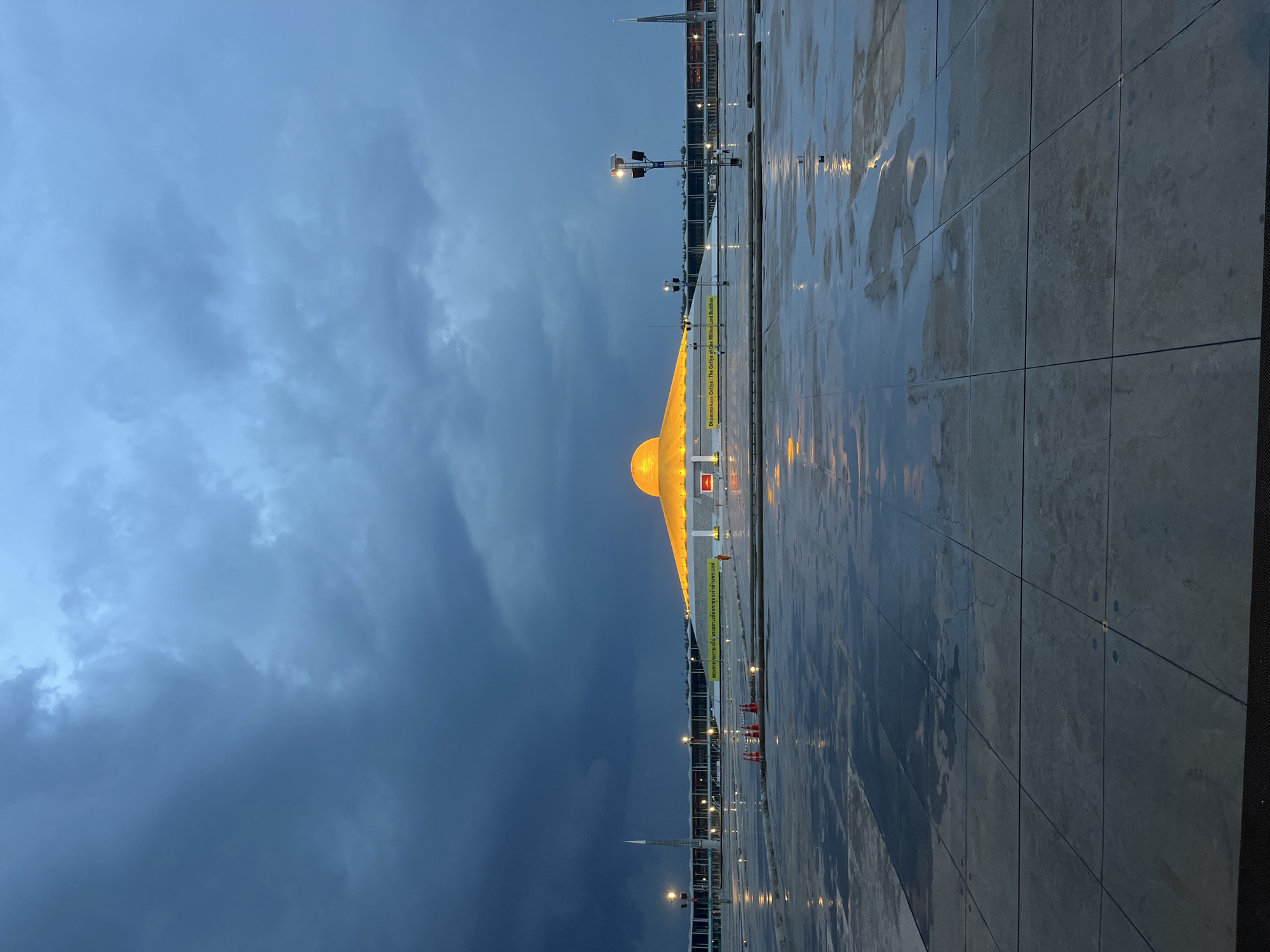 The image depicts the Dhammakaya Cetiya, also known as The Cetiya of One Million Lord Buddhas, located in Tha Khlong Town Municipality, Thailand. The structure is a large, golden dome illuminated from within, set against a dramatic sky filled with dark, turbulent clouds. The foreground consists of a vast, wet, tiled plaza, reflecting the ambient light from the structure and street lamps. Several traffic cones are visible on the plaza. In the background, a tall, slender spire and a long, low building with lights are visible. The overall atmosphere is moody and overcast, suggesting imminent or recent rainfall.