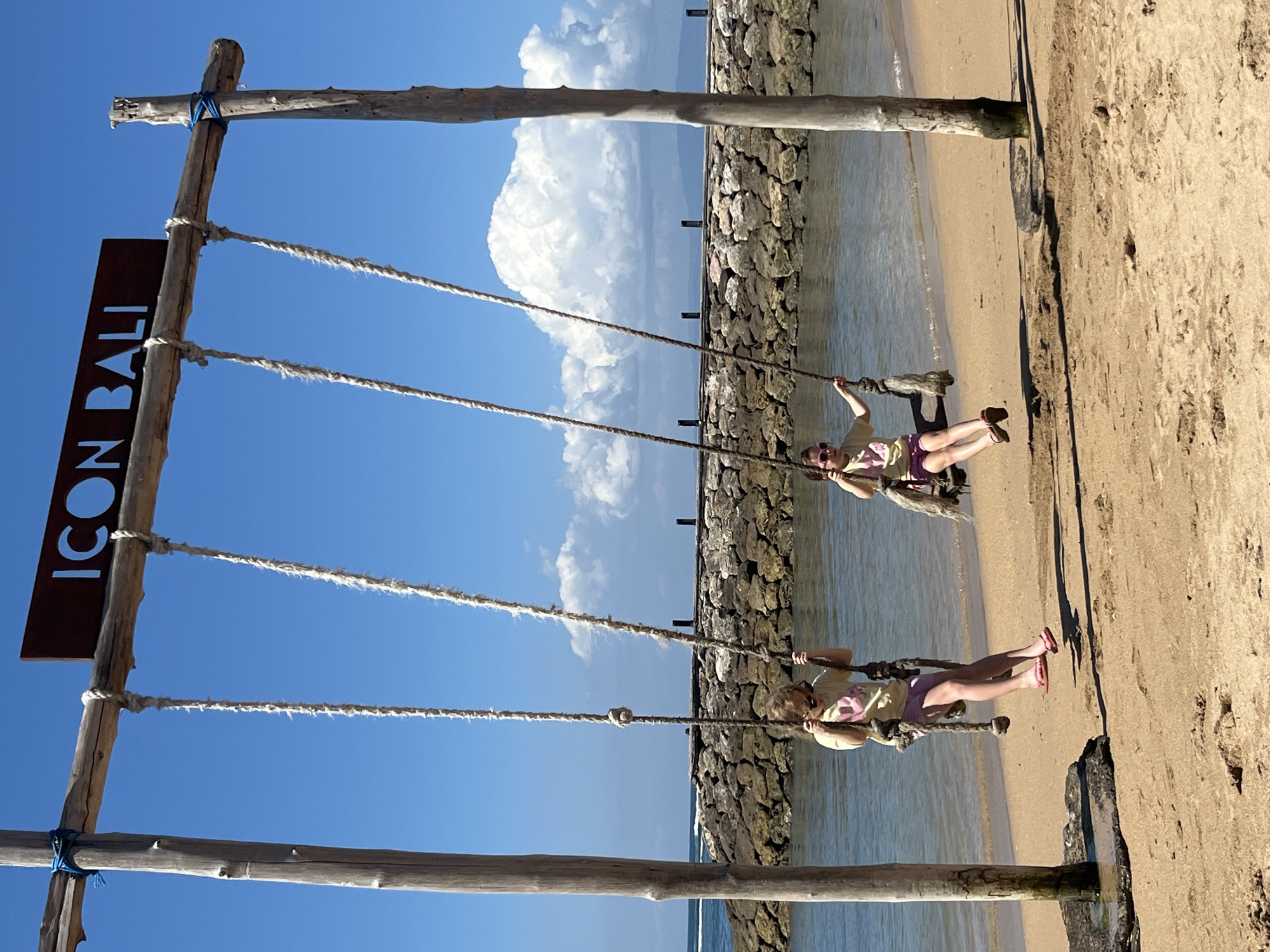 Two young girls are on swings at a beach location in Denpasar, Indonesia. The swings are suspended from a large wooden frame structure. One girl is wearing a yellow t-shirt and purple shorts, and the other is wearing a similar outfit. They are both looking forward. The beach has sand and the ocean is visible in the background. There is a stone wall or breakwater in the distance, and some pilings are visible in the water. The sky is blue with some white clouds. A sign with the word "ICON BALI" is attached to the wooden frame above the swings.
