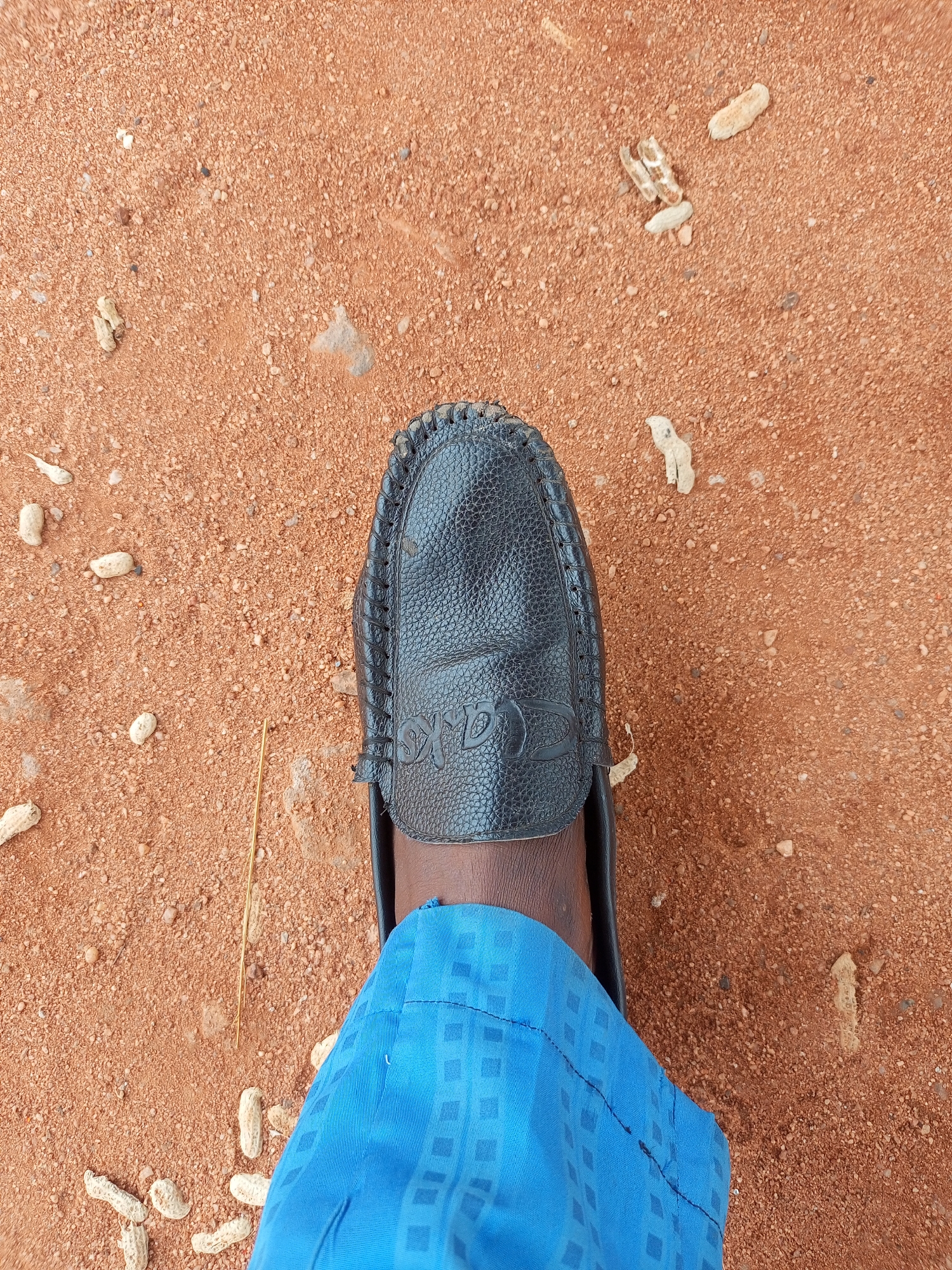 The image is a close-up, top-down view of a person's left foot wearing a black leather loafer and blue patterned pants, standing on a reddish-brown dirt surface. The dirt is composed of fine particles and small pebbles, with several peanut shells scattered across it. A thin, dry piece of grass or straw lies diagonally across the dirt near the shoe. The loafer is textured and has a stitched trim around the opening. Embossed on the surface of the shoe are the letters "AXS" in a stylized font. The blue pants have a repeating geometric pattern of darker blue squares. The scene appears to be outdoors, potentially in a rural or less developed urban area, given the natural ground cover and scattered debris. No specific landmarks or clues point to the city of Tempure, Nigeria.