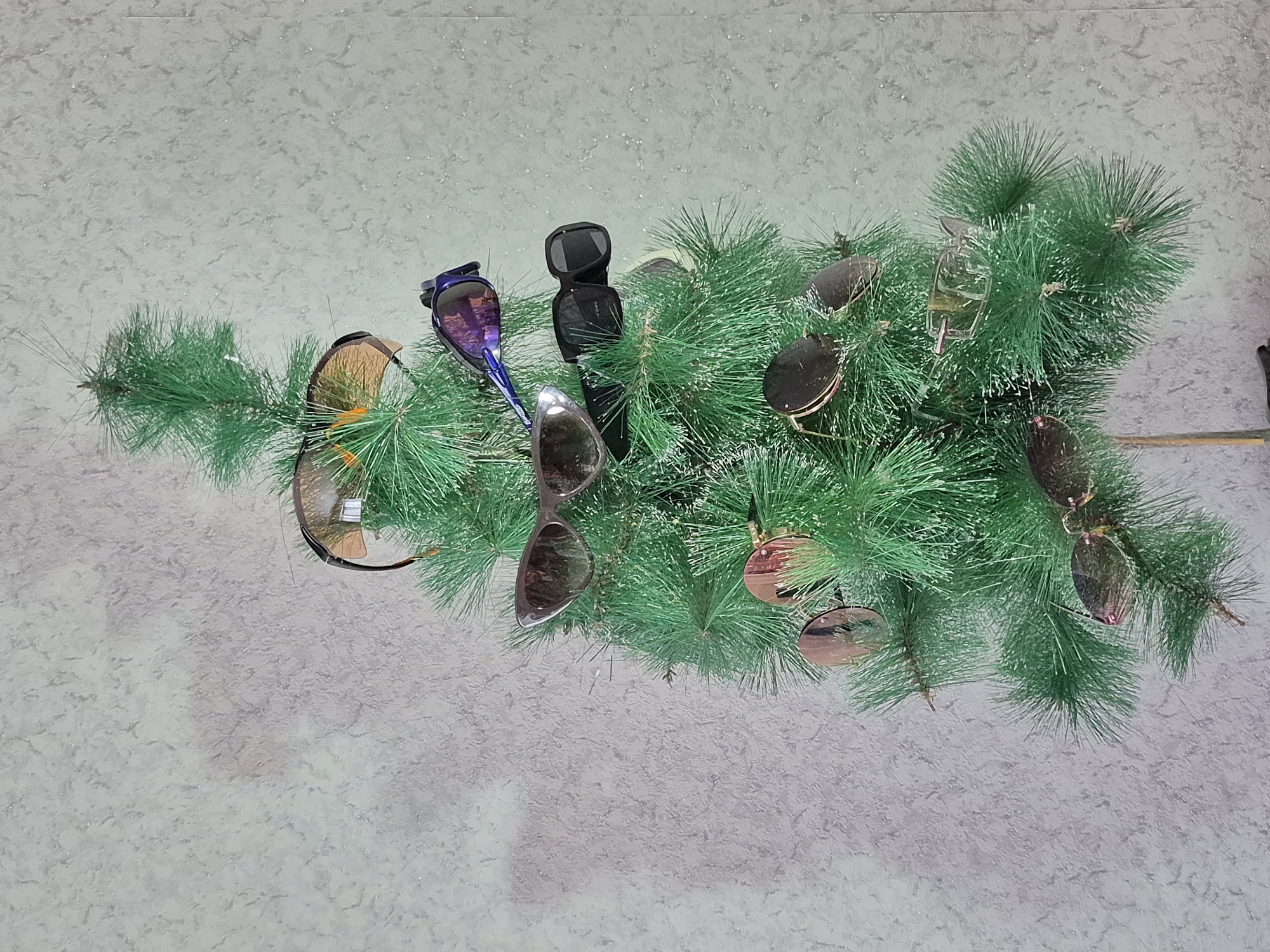 The image shows an artificial pine branch laid on a textured gray surface, possibly a countertop or floor. Several pairs of sunglasses are placed amongst the pine needles, appearing as decorations or ornaments. The sunglasses vary in style, color, and size, including black, brown, blue, and silver frames. The overall scene gives a casual, perhaps playful, impression, but the location is indeterminate and not in the city of Unknown, Unknown.