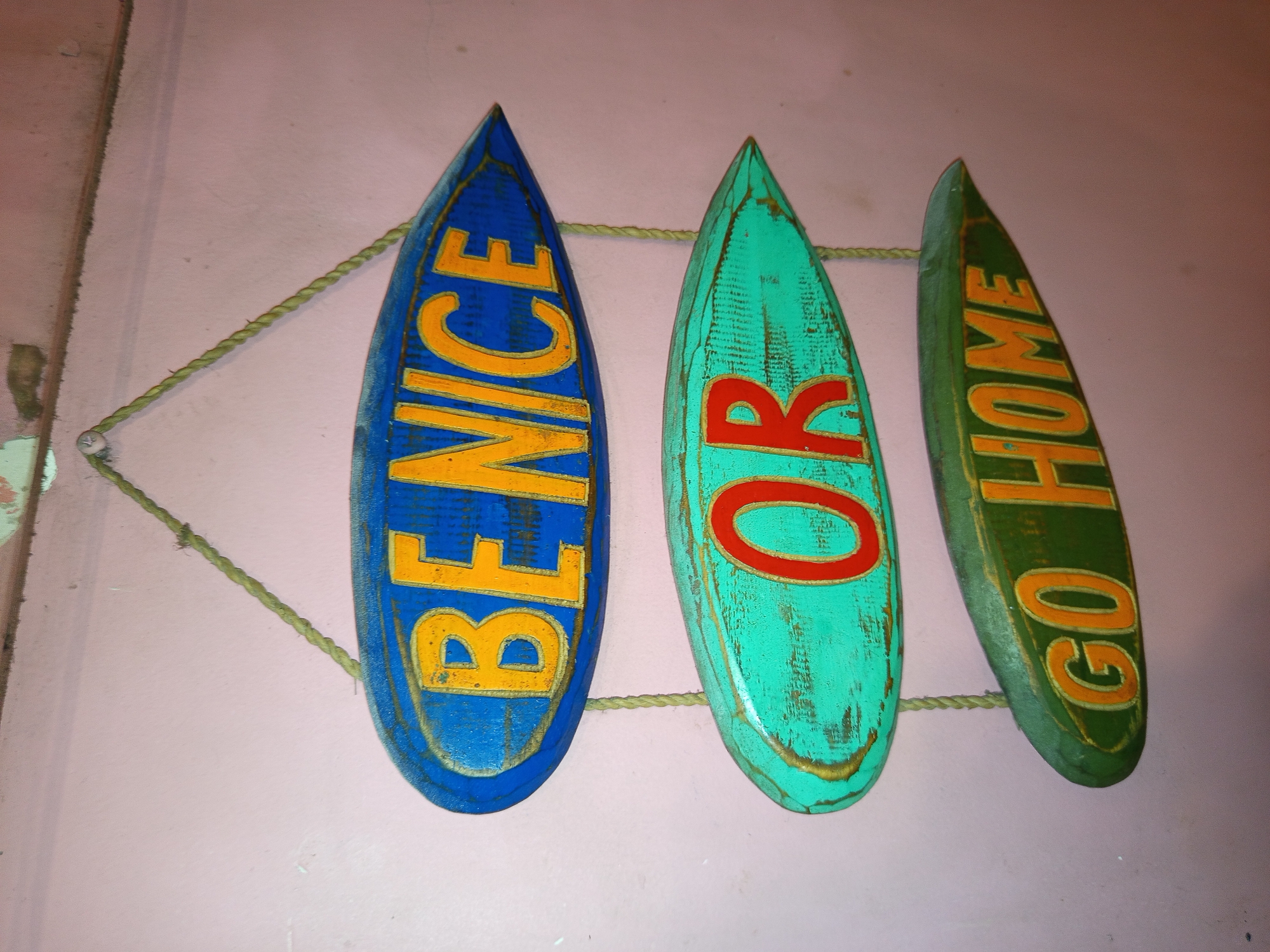 A close-up photograph shows three surfboards hanging on a light pink wall in an unidentified location, possibly in North Kuta, Indonesia. Each surfboard is a different color: the leftmost one is blue, the center one is teal, and the rightmost one is green. The surfboards are oriented vertically and arranged horizontally, slightly angled. Each board has a word written vertically on it in a contrasting color. The blue board has the word "BE NICE" written in yellow, the teal board has the word "OR" in red, and the green board has the words "GO HOME" in yellow. The boards are hung together by a length of light-colored rope. The texture of the surfboards appears distressed or weathered, suggesting they are vintage or have been given a rustic appearance. The background is a light pink wall with an uneven texture.