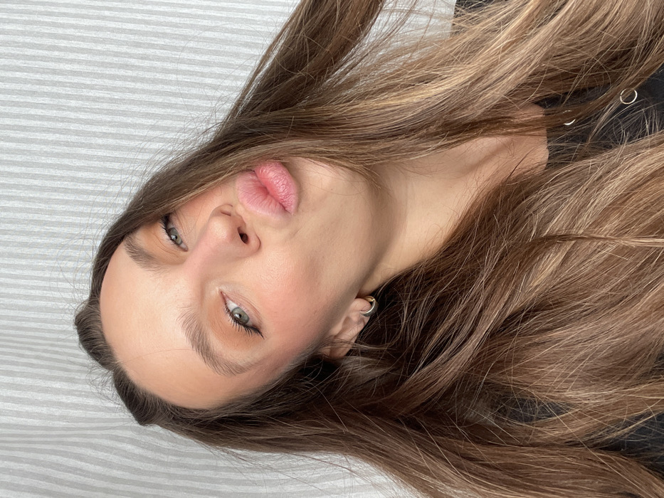 The image displays a close-up, head-and-shoulders portrait of a female individual. She possesses long, brown hair with lighter highlights, styled to frame her face. Her light-colored eyes are directed towards the camera, and her lips are pursed in a kiss-like gesture. Facial features include defined eyebrows, visible blush on the cheeks, and eye makeup. A single gold-colored hoop earring is present in her left ear. She is clothed in a dark garment, portions of which are visible at the lower edge of the frame, exhibiting small metallic circular details. The background is a uniformly light-toned surface featuring distinct vertical stripes, consistent with an interior wall or textile. The specific location within Budva, Montenegro, cannot be ascertained from the visual evidence.
