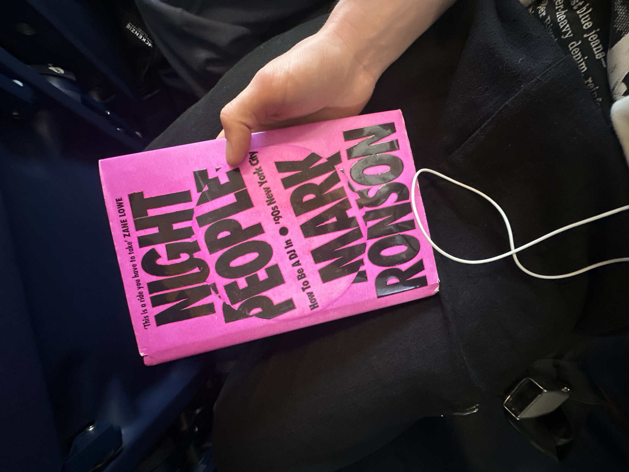 A close-up, slightly high-angle shot shows a person holding a bright pink book, resting on their lap which is covered in black clothing, while seated. A pale hand grips the book on the left side. A white earbud cable trails from the right side of the book across the person's black-clad lap.
The book's prominent pink cover displays bold black text: "NIGHT PEOPLE" and "MARK RONSON". Below "MARK RONSON", smaller text reads "How To Be A DJ In '90s New York City". A quote, "'This is a ride you have to take' ZANE LOWE", is printed along the left edge of the cover.
The person is wearing black clothing, possibly a hoodie or jacket, which has some partially visible white text. Readable fragments include "ACKENZIE" (likely part of a brand name "MACKENZIE") and reversed text such as "tblue jeans-", "erheavy denim, reinfor", and "Per Riv". A silver seatbelt buckle is visible against the black fabric in the lower right of the frame, indicating the person is in a vehicle. The background features a blurred dark blue surface, likely part of a seat or vehicle interior. The scene suggests the person is reading or listening to audio while traveling, possibly within Uttlesford, United Kingdom.