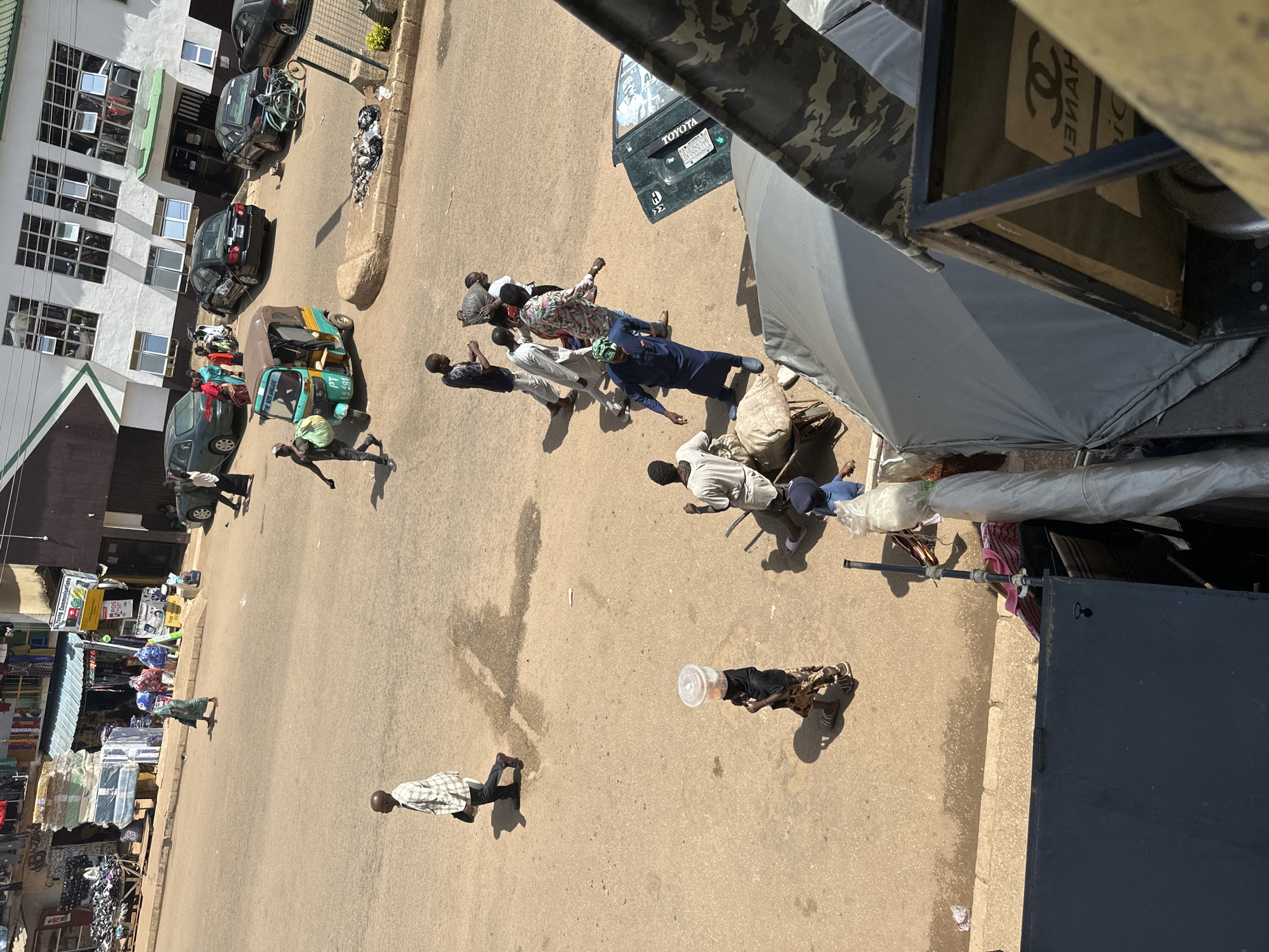 This media file captures a bustling street scene in Bukuru, Nigeria, likely during daylight hours given the strong shadows. The setting appears to be a marketplace or a busy urban thoroughfare, with various shops and buildings lining the street. Several people are engaged in activities, suggesting a normal day of commerce and movement.
In the foreground, a person in a plaid shirt walks across the road, casting a long shadow. Further down the street, another individual wearing a black garment and a patterned skirt is walking with a clear plastic container on their head. Other people are seen walking and interacting, with some carrying bags and others seemingly conversing. A colorful auto-rickshaw, often used for public transport in Nigeria, is parked on the side of the road. Several cars, including what appears to be a Toyota, are also visible, indicating vehicular traffic.
The scene is lively, with a sense of everyday activity. The architecture of the buildings suggests a tropical or subtropical climate, and the bright sunlight indicates fair weather. Some of the shops display signs with text, though much of it is too small or blurry to decipher. One sign on a vehicle clearly reads "TOYOTA" and has a license plate. Another sign on a shopping bag has the Chanel logo visible. The overall impression is one of a vibrant community going about its daily life.