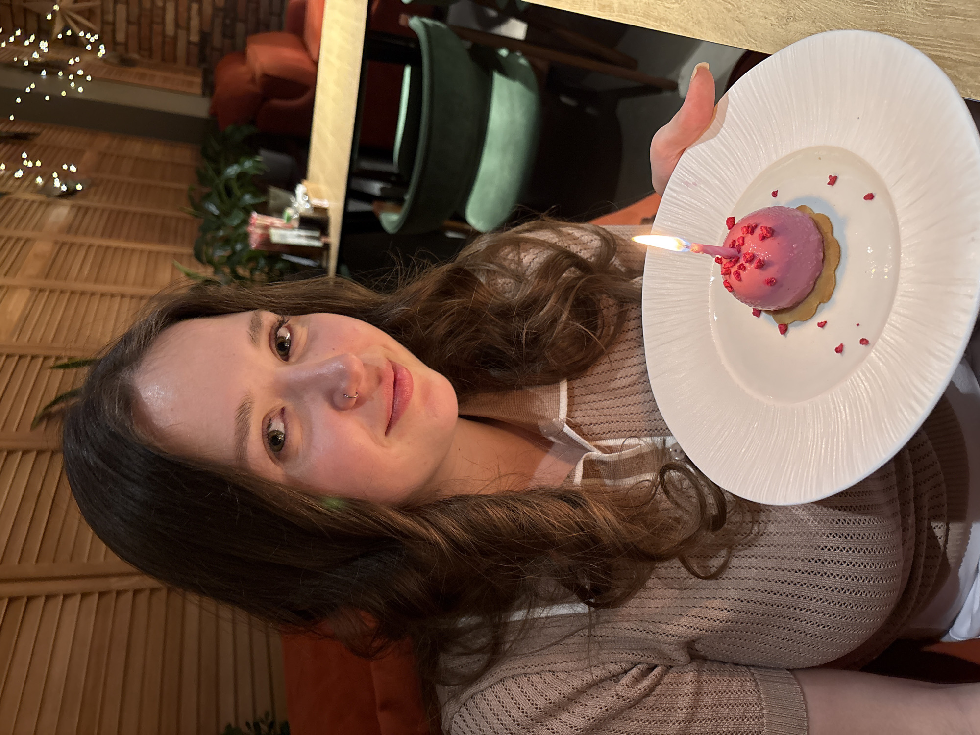 The image, rotated 90 degrees clockwise, features a young woman holding a celebratory dessert with a lit candle. She occupies the foreground, smiling gently and looking directly at the camera. She has long, wavy brown hair, light skin, brown eyes, and a nose piercing. Her attire includes a light brown, open-knit cardigan or top worn over a lighter undergarment.
In her right hand, she presents a white plate with a spherical, pink dessert, possibly a mousse or small cake, garnished with red sprinkles. A lit pink candle with a blue flame is embedded in the dessert, indicating a birthday or other special occasion. The plate has a subtle ridged texture.
The setting is an indoor space, likely a restaurant or cafe in Izhevsk, Russia, characterized by a warm and inviting ambiance. The background reveals wooden slatted walls, creating a cozy texture. Out-of-focus decorative string lights sparkle on the left, while upholstered chairs in deep green and burnt orange hues are visible further back. The lighting is soft and warm, contributing to the festive mood. The woman's expression conveys happiness and contentment, reflecting the celebratory event. No visible text is present in the image.