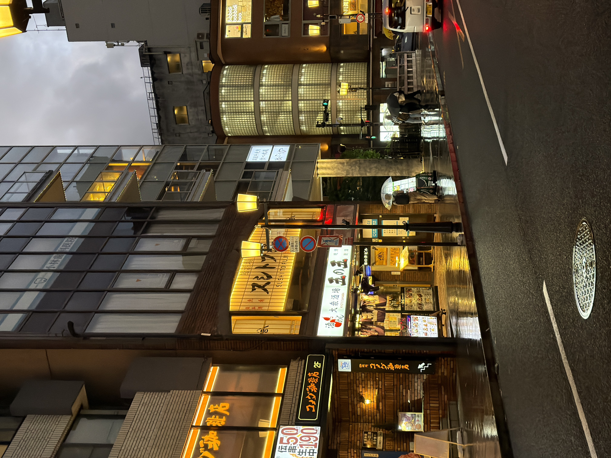 This image depicts an urban street scene at dusk or night, wet from rain, in the Kawaramachi area of Kyoto, Japan. The specific location is identifiable by visible signage as the vicinity of 284-5 Shijocho, Shimogyo Ward, Kyoto, 600-8002, Japan. The foreground features a wet asphalt road with a faint white lane divider line. A circular, ornate metal manhole cover is visible in the lower center. The wet road and sidewalk surfaces intensely reflect the artificial lights from the surrounding buildings and street furniture. In the mid-ground, on the left sidewalk, a single individual stands under a transparent umbrella, facing away from the viewer, next to an illuminated white menu board. Further right, closer to the center of the image, three individuals are gathered; two are positioned under a dark umbrella, with one person bent over, seemingly attending to or interacting with something low to the ground. A white commercial van with illuminated red taillights is visible on the right side of the road, positioned in traffic and facing away. Several yellow-lantern streetlights illuminate the scene. A traffic light pole displays a green signal facing the viewer. Various Japanese street signs are mounted on poles, including regulatory signs such as a blue circle with a red diagonal line (indicating no stopping or parking) and a red circle with a blue bar (indicating a restriction or prohibition). The background is dominated by multi-story commercial buildings densely packed along the street. On the far left, a building with a brick-patterned lower facade and large upper-story windows displays prominent yellow and orange neon signs. Legible signs include "コメダ珈琲店" (Komeda Coffee Shop) and "串かつ伝串" (Kushikatsu Denshaku), with pricing "50/190" also visible. Adjacent to this, a taller building features multiple large windows, with "小林歯科医院" (Kobayashi Dental Clinic) visible on a sign through a reflection or window. Further right, a distinctive multi-story, cylindrical architectural element constructed entirely from rectangular glass blocks is prominently illuminated from within, casting a geometric grid pattern. To the right of the glass-block structure, another building section has multiple windows glowing with yellow light, and a partial sign reading "Bibbidi bobbidi boo" is visible. Below this section, a large, brightly lit sign reads "酒と大衆酒場 玉の玉" (Sake and Public Izakaya Tamanotama) with "KYOTO SAKABA" in English below it. Above this, another illuminated sign displays "スシトフシ" (Sushi Tofuji) and "SUSHI TOFUJI" in English. Another sign on a gray building in the upper right background