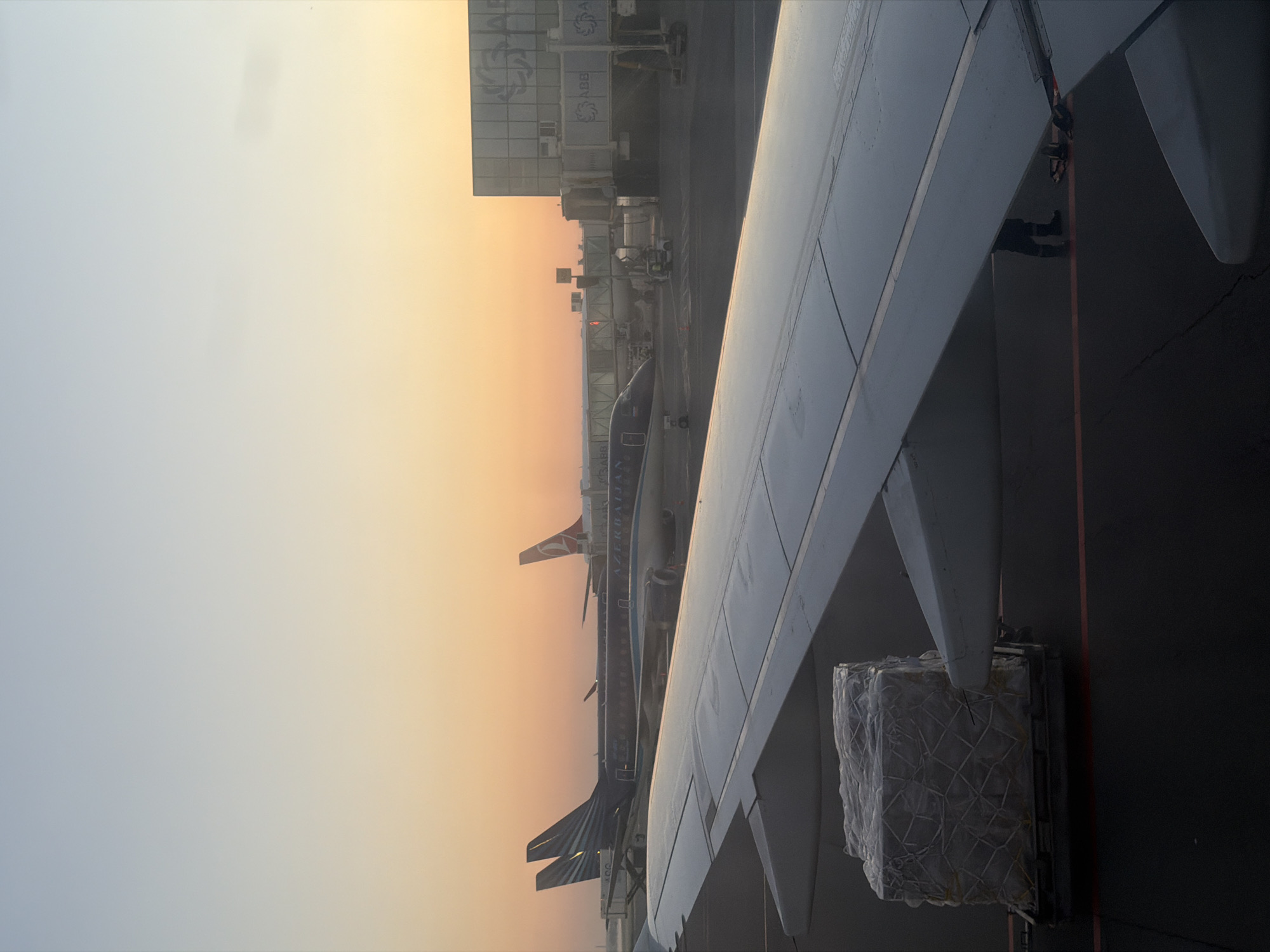 The image depicts an airport tarmac at sunrise, with the warm hues of the sky fading from orange to a soft blue. In the foreground, the wing of an aircraft is prominent, extending diagonally across the frame. A cargo container, wrapped in protective netting, is situated on a pallet and positioned on the tarmac adjacent to the wing.
Beyond the wing, several commercial airplanes are parked at gates, connected by jet bridges. One aircraft, bearing the livery of Azerbaijan Airlines, is clearly visible. The airport terminal buildings and other airport infrastructure are present in the background. The scene appears to be at Heydar Aliyev International Airport, located in the Khazar Raion of Baku, Azerbaijan. The overall atmosphere is one of early morning activity at an international airport.