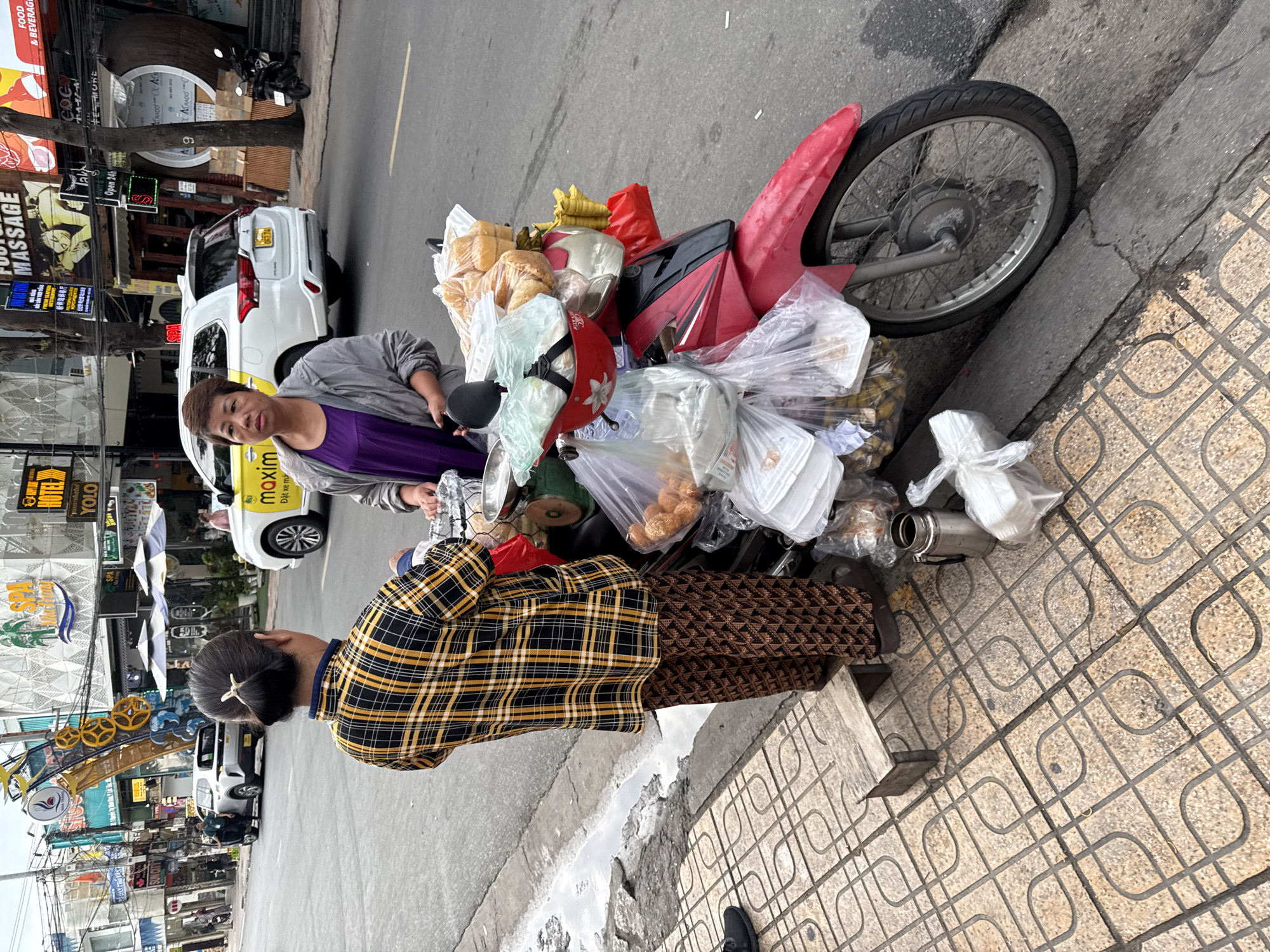 This image depicts a street scene in Nha Trang, Vietnam, where two women are interacting with a food vending motorcycle. The woman on the left, wearing a yellow and black plaid shirt and patterned brown pants, stands on a tiled sidewalk. She appears to be in conversation with the woman on the right. The woman on the right, dressed in a purple shirt and gray jacket, is positioned behind the motorcycle.
The motorcycle, which is red and white, is heavily laden with food items packaged in plastic bags and containers. Prominently visible are loaves of bread and what appear to be fried snacks. A green scale is also mounted on the motorcycle, suggesting it's part of the vending setup.
The background reveals a typical urban street environment in Nha Trang. Buildings with signs for businesses like "SPA," "HOTEL," and "FOOT MASSAGE" line the street. A white car with yellow accents and the word "MAXIM" is parked nearby, indicating a potential transportation service. Other vehicles and pedestrians are present but less prominent. The street is paved, and a curb separates the roadway from the sidewalk. The weather appears overcast. The overall scene captures a moment of daily commerce on a bustling Vietnamese street.