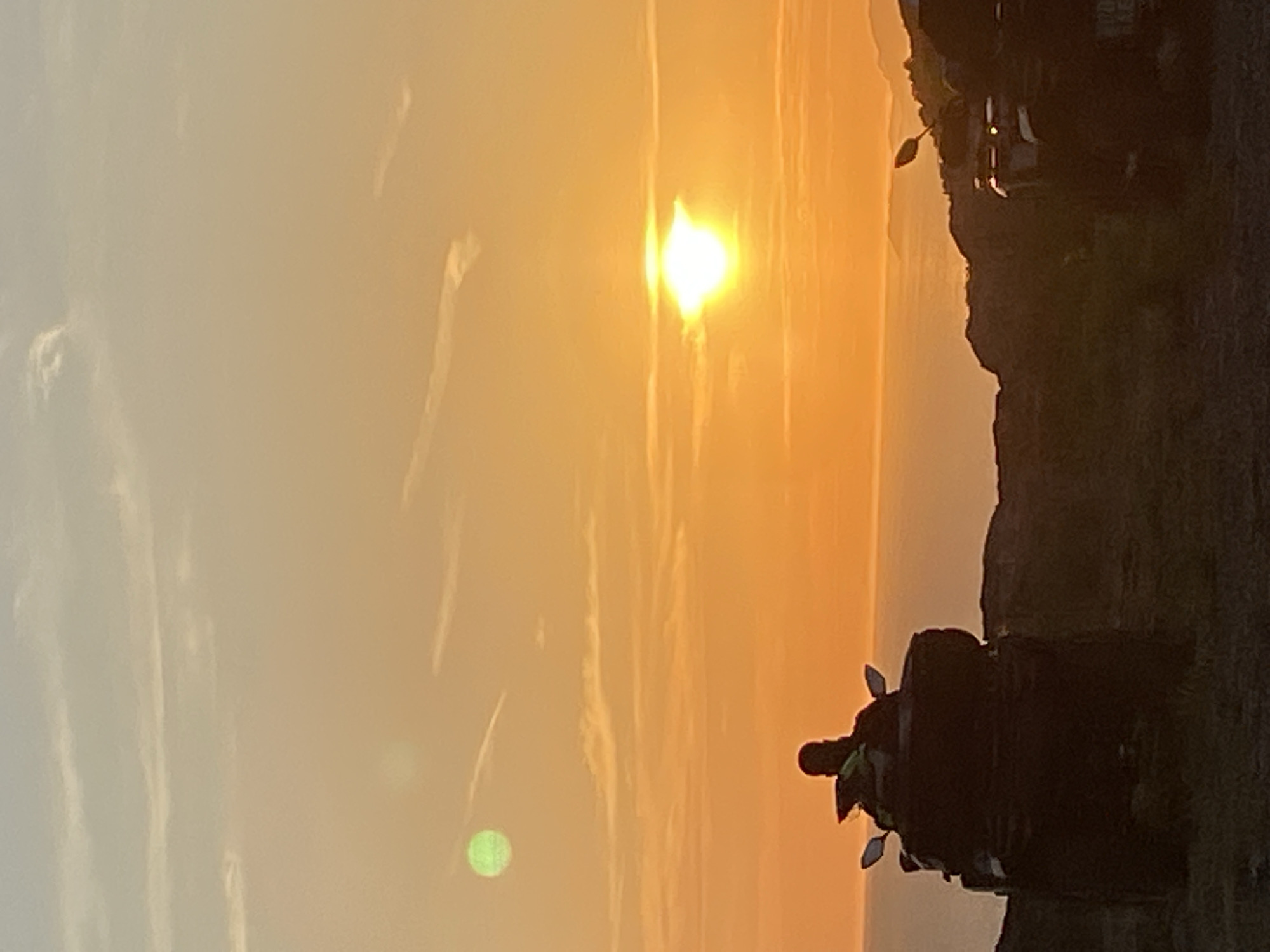 A man on a dark ATV faces away from the camera, looking towards the ocean at sunset. The sun is low on the horizon, casting a warm orange glow across the sky and water. The ATV is parked on a dirt path, with another ATV visible in the background. The scene appears to be set in a coastal area, possibly Montenegro, given the mention of Kovačko Polje. The sky is partly cloudy, with wispy clouds catching the sunlight. There is a hint of green light in the upper left quadrant of the image, possibly a lens flare.