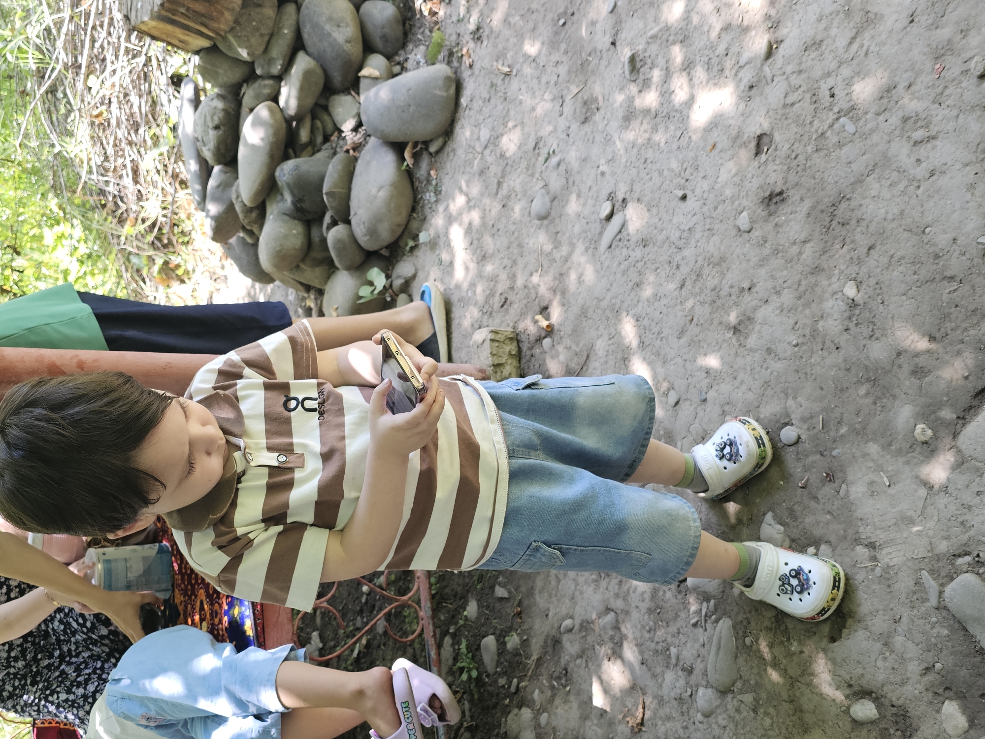 The image shows a young boy standing on a dirt path in a park or outdoor area, possibly near a riverbed due to the presence of large stones. The boy is wearing a brown and white striped polo shirt, denim shorts, green socks, and white clogs with car decals. He is holding a smartphone in his hands and looking down at it. In the background, a pile of smooth, gray river stones is visible, along with some dry brush and foliage. To the left, another child's legs are visible, wearing light blue shorts and pink sandals. A person's arm, wearing a dark floral patterned dress, is also partially visible in the background, holding a light blue container. The setting appears to be a natural, somewhat untamed environment with dappled sunlight filtering through trees. The location is identified as Ден-Булак 2, Kyrgyzstan.