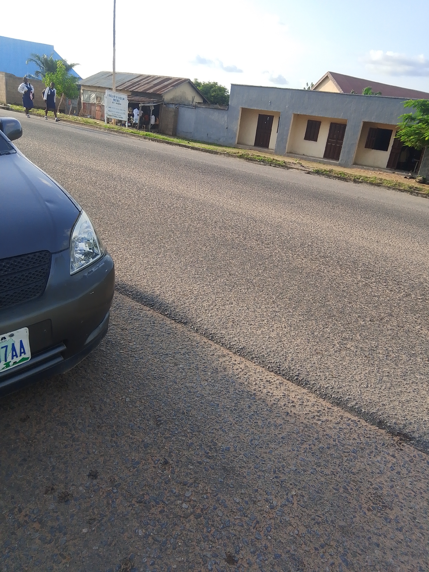 The image depicts a street scene in Jalingo, Nigeria, likely during the daytime under fair weather conditions, as indicated by the clear sky and shadows. In the foreground, the front of a dark-colored car is visible, facing left. Its license plate reads "37AA".
Across the road, several structures line the street. To the left, two individuals in school uniforms, appearing to be female students, walk on the sidewalk. Behind them, a building with a blue roof is partially visible. Adjacent to this is a small shop with a corrugated metal roof, where several people are gathered. A sign above the shop displays text, but it is partially obscured and difficult to read in full.
Further down the street, a row of connected buildings with grey facades and cream-colored sections are visible. These buildings have several doorways and windows, suggesting they might be shops or offices. The road itself is paved with asphalt and shows a clear division line. The overall impression is of a typical urban or semi-urban environment in Nigeria, with daily life unfolding on the street.