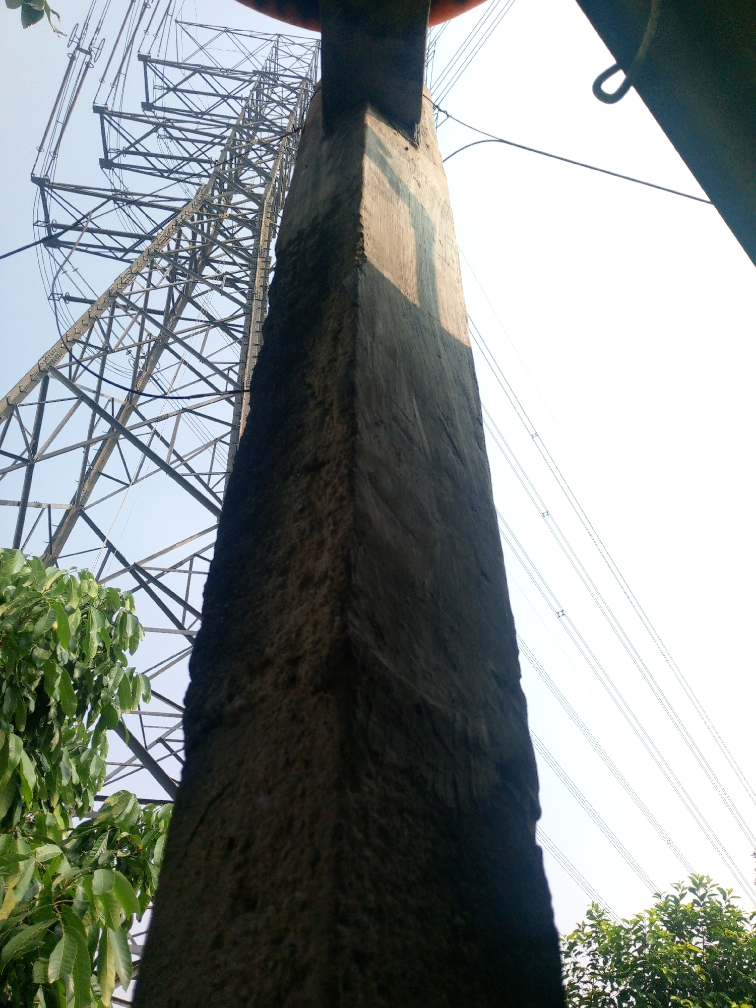 The image shows a low-angle view of a tall electricity pylon against a clear sky. In the foreground, a rough concrete pillar runs vertically, obscuring a portion of the pylon. To the left of the pillar, lush green foliage from a tree is visible. Multiple power lines stretch across the sky behind the pylon. The scene suggests an outdoor industrial or infrastructural area. The location is identified as Dukuh, Indonesia.