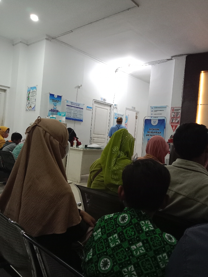 The image depicts an indoor waiting area, likely a clinic or public service office, in Gladaksari, Indonesia. The scene is illuminated by overhead fluorescent lighting from a white ceiling. Multiple individuals are seated, facing away from the camera, indicating they are waiting.
In the immediate foreground, a person, possibly a child or young male, is seated with their back to the camera, wearing a dark green shirt with a light green geometric pattern. To their left, another individual wears a light brown hijab and a white face mask, also facing away. Further left, partially visible, is a person in a yellow hijab. Behind the person in the brown hijab, a individual in a light green hijab with subtle embroidered patterns is seated. Another seated person in a reddish-brown hijab is visible further right, behind the person in the green hijab. A male adult in a light brown/beige collared shirt is seated behind the person in the patterned green shirt.
In the mid-ground, a male individual in a light blue shirt and a light-colored hat (possibly a traditional 'peci') stands facing a light-colored counter or desk, appearing to interact with an unseen person or service point. Behind this counter, two white closed doors are visible.
The white walls are adorned with various informational posters and signs. From left to right, a poster with blue and green graphics, a blue poster with text, a larger blue poster displaying a schedule or list, and a blue banner on a stand stating "MOHON BERSABAR MENUNGGU" (Please be patient while waiting) in Indonesian. A red fire extinguisher is mounted on the wall near this banner. The seating consists of multiple rows of chairs with dark, perforated seats and backs, set within metal frames. A dark column is visible on the far right of the frame.
