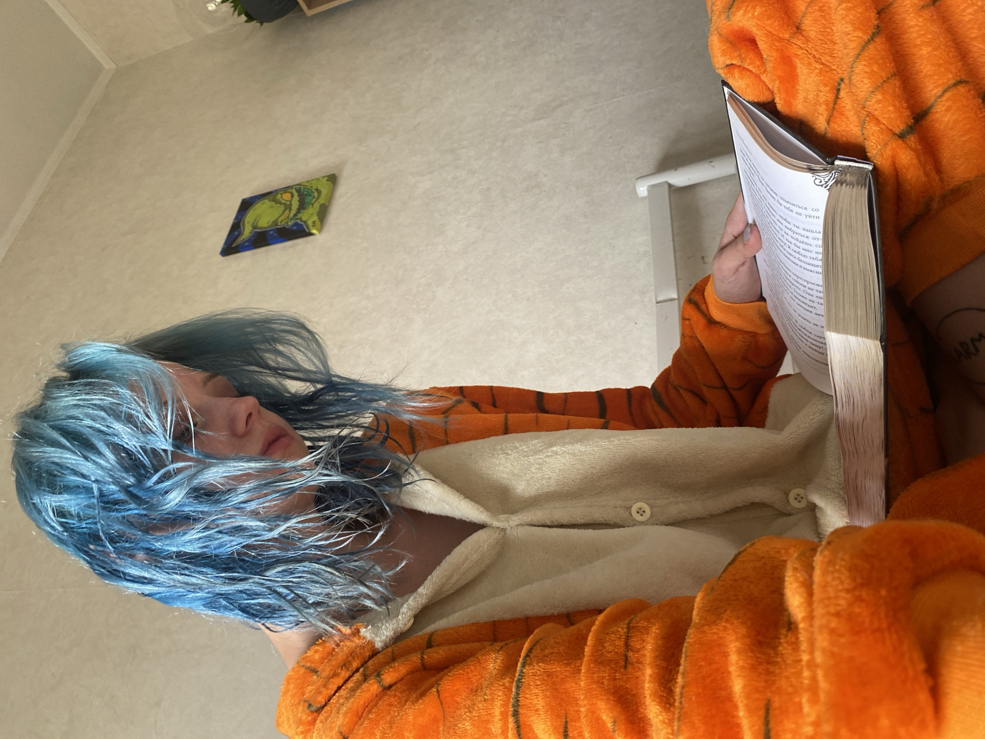 A person with bright blue hair, wearing an orange and black tiger-striped onesie, sits indoors and reads a book. The person's head is tilted down, focusing on the book, and their expression is one of concentration. The onesie is fluffy and bright orange with black stripes, typical of a character costume. The book is open and appears to be a paperback with a significant number of pages. In the background, a textured wall with a framed picture of a green, cartoonish monster is visible. The room appears to be a bedroom or living space, with soft lighting. There is no indication of the location being in Bishkek City, Kyrgyzstan.
