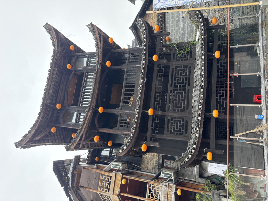 This image captures a multi-story traditional Chinese wooden building located in Furong, China, under an overcast sky. The central structure is dark-stained wood, featuring three visible levels, each characterized by layered, curved, tiled roofs and balconies with intricate wooden lattice-work railings. Approximately twenty-four orange, spherical lanterns are suspended from the eaves and railings of this main building. To its left, a lighter-colored wooden building with similar architectural elements, including balconies and carved railings, is partially visible, displaying two external air conditioning units and a few yellow lanterns. On the right, a segment of a stone wall and a vertical bamboo pole draped with a blue and white patterned fabric are present. The immediate foreground features a modern glass and metal railing. Behind this barrier, on the ground, are several brooms leaning against a wall, a red plastic stool, a blue container, and two rolls of white material. No individuals are present in the scene.