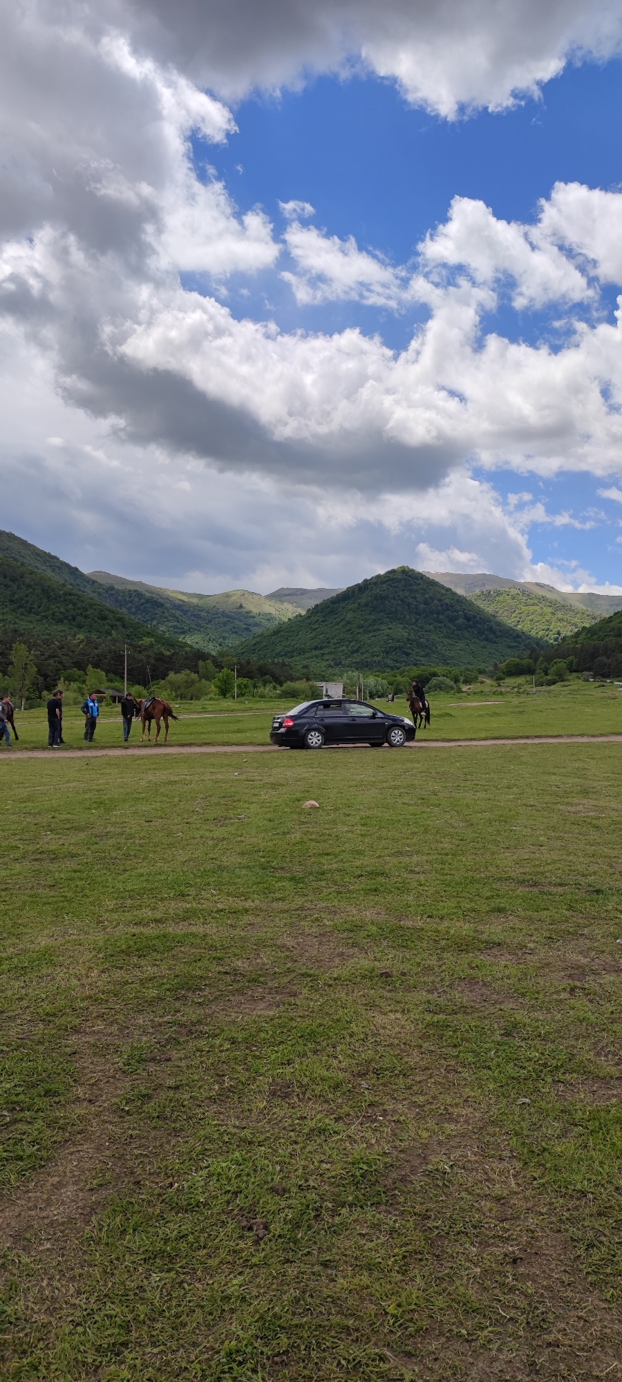 The image depicts a rural scene in Gyulagarak, Armenia, featuring a black sedan positioned on a dirt road that bisects a large green field. A group of people stands to the left of the car, some near a brown horse. Another individual is mounted on a horse to the right of the car. The background showcases rolling green mountains partially obscured by a sky filled with puffy white and grey clouds.