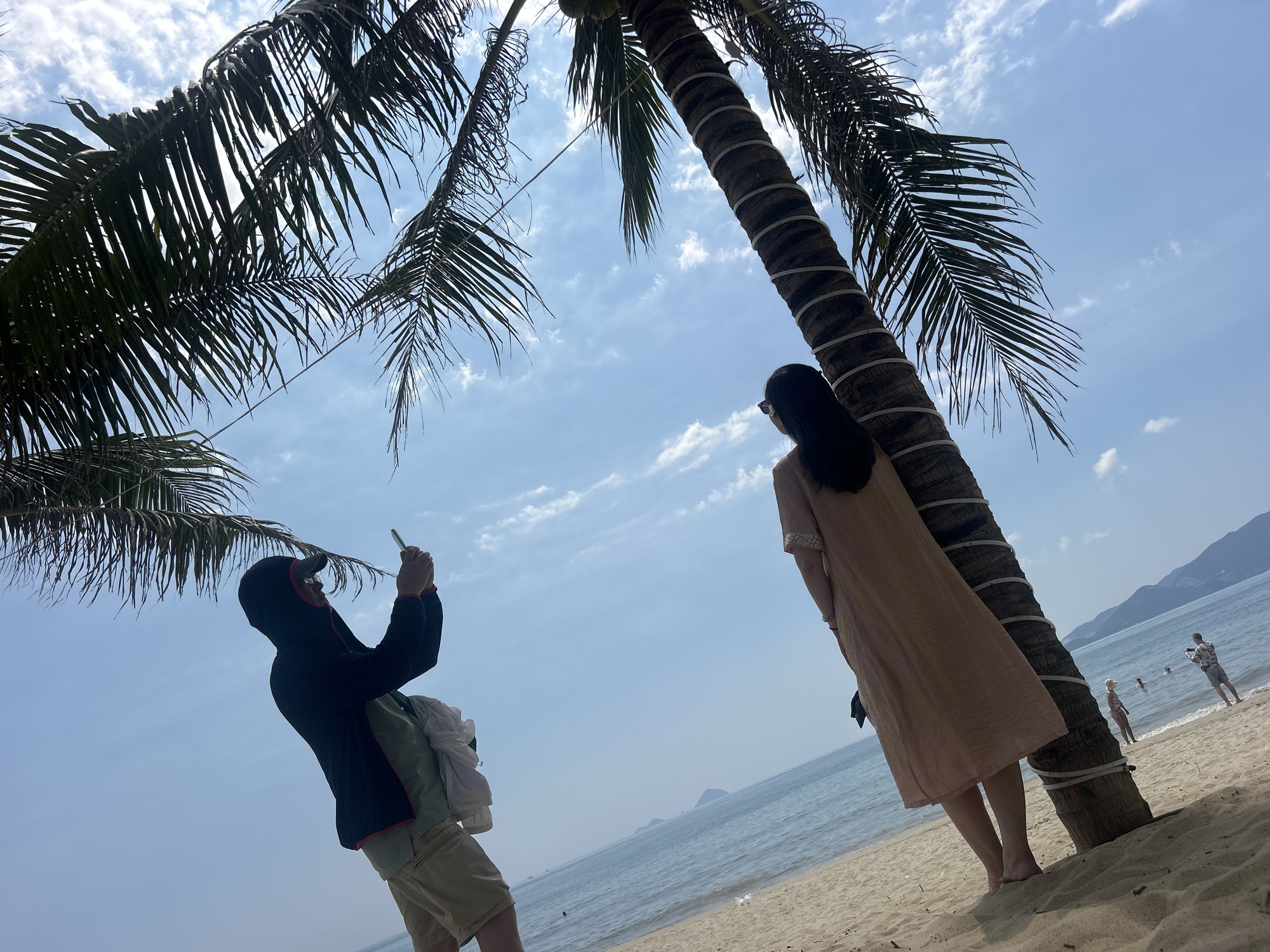 A medium shot captures a beach scene in Nha Trang, Vietnam. A slender woman with long dark hair and sunglasses, wearing a pale pink knee-length dress, stands barefoot facing the ocean, leaning slightly against the trunk of a decorated palm tree. A man, dressed in a dark hooded jacket, khaki shorts, and a light green shirt, stands at an angle to the woman, holding up a phone as if taking a picture. In the background, a stretch of light brown sand leads to a calm, blue ocean. Further out, a few individuals can be seen wading in the water. In the distance, a mountainous island is visible. The sky overhead is pale blue with wisps of white clouds, partially obscured by the fronds of the palm trees. The palm tree has white lights wound around its trunk.