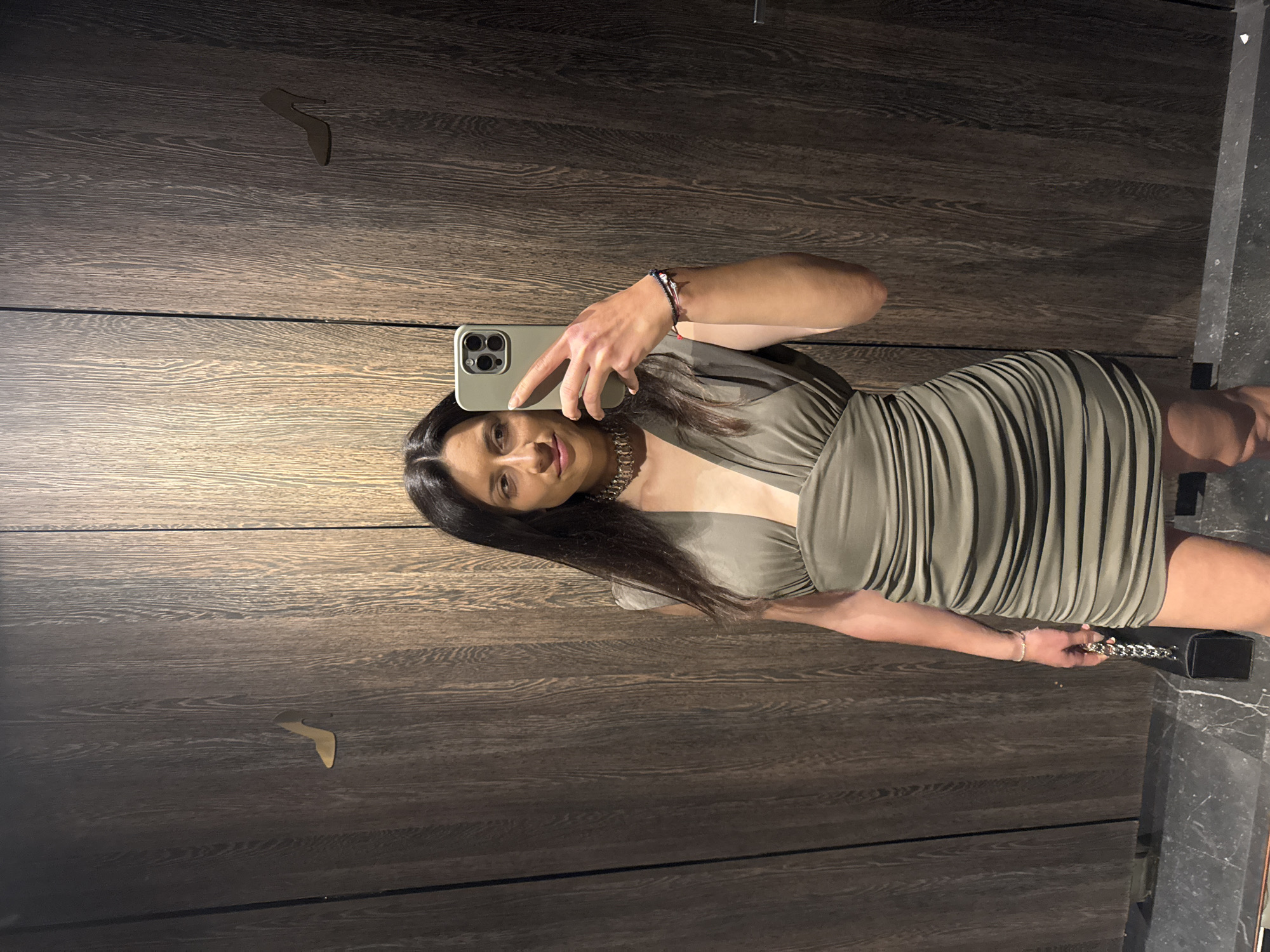 A woman is taking a selfie in a room with dark wood paneling on the walls. The woman is wearing a short, olive-green dress with a low neckline. She is holding a phone in her left hand and a black purse with a chain strap in her right hand. The phone has a light green case. The room appears to be a fitting room or dressing area, possibly in a store or shopping center in Blagoevgrad, Bulgaria, though there is no specific detail to confirm this location.