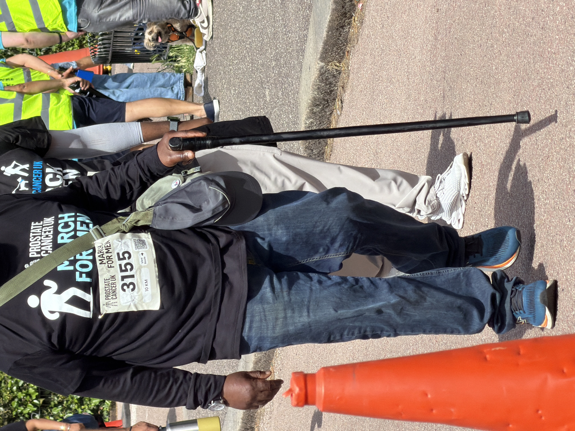 The image shows a close-up of a man participating in the "March for Men" event organized by Prostate Cancer UK. He is wearing a black long-sleeved t-shirt with the event's logo and a race bib with the number 3555. The man is walking with the aid of a black cane and is wearing blue jeans and athletic shoes. In the background, other participants, some wearing high-visibility vests, are visible, along with a small dog on a leash. The event appears to be taking place outdoors, on a paved surface, likely in a public area. The presence of an orange traffic cone suggests a possible organized route or a controlled environment. The overall scene depicts an event for a charitable cause, emphasizing community participation and support.