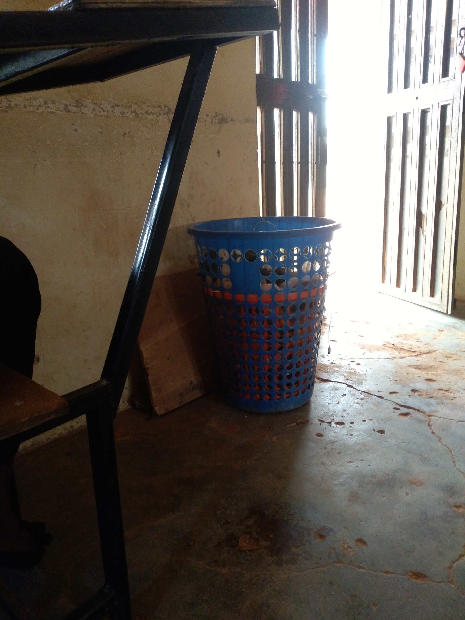 The image depicts an interior scene, likely in the city of Jos, Nigeria. In the foreground, a blue plastic laundry basket with orange contents is prominently placed on a concrete floor. The basket is cylindrical and features a perforated pattern. To the left, a black metal table structure with a wooden surface is visible, partially obscuring the background. Behind the basket, a piece of light brown cardboard leans against a wall. The wall itself is a textured, light brown color with visible wear and tear. In the background, a doorway with vertical white bars leads to a brightly lit outdoor area, suggesting daylight. The floor exhibits a rough texture with several reddish-brown stains and cracks, indicating it is likely a well-used and possibly older surface. The overall lighting suggests a poorly lit interior contrasted with a strong light source from the exterior.