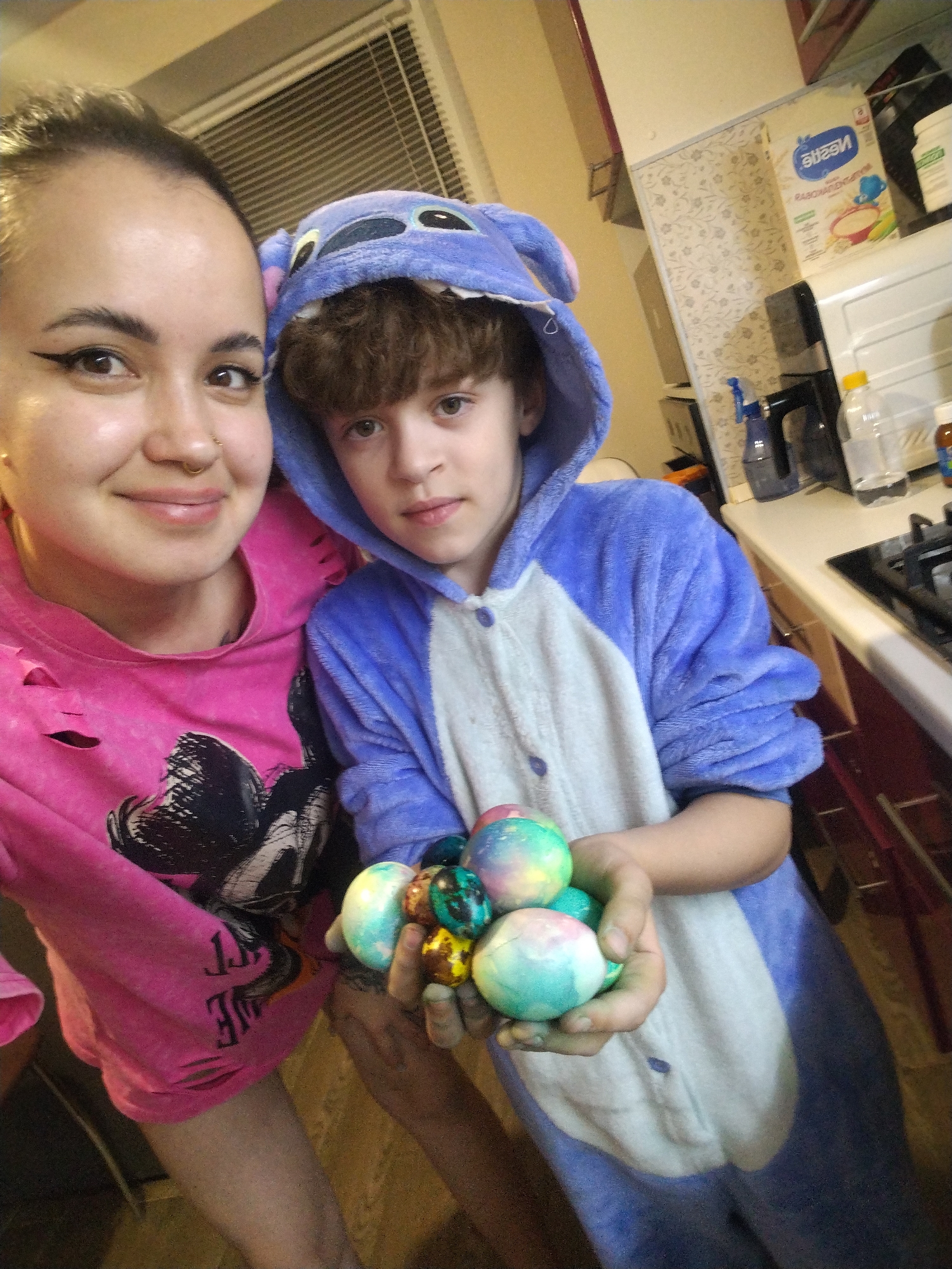 A selfie taken indoors features a woman and a child. The woman, positioned to the left, has her hair pulled back and is wearing a pink, distressed hoodie with a black graphic. To her right, the child is wearing a blue and white onesie resembling a cartoon character. The child holds a cluster of decorated Easter eggs. In the background is a window with blinds, a kitchen counter with various items, and wallpaper. The scene does not provide enough information to identify a specific location in the city of Noviy, Russia.