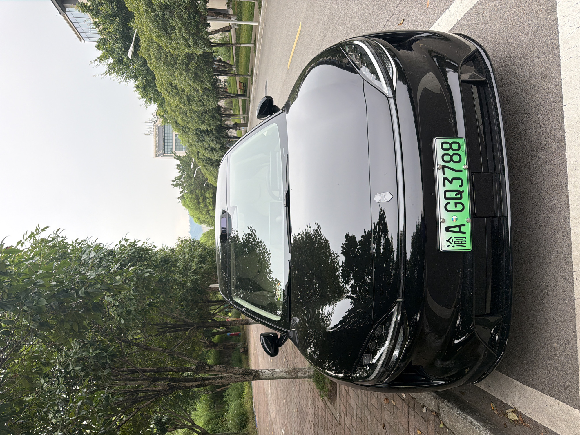 A black electric car, identified by its license plate "渝A GQ3788", is parked on a street in Jiangjin, China. The car is a sedan with a sleek, modern design, featuring LED headlights and a prominent sensor array on the roof. The vehicle is positioned at an angle on the right side of the road, with its front facing the camera. The street is lined with trees on both sides, and in the background, there are buildings and a hazy sky. The car is parked next to a sidewalk made of brick pavers, and there is a curb between the sidewalk and the road. The road itself is paved and marked with white lines and a yellow line. There are no people visible in the image.
