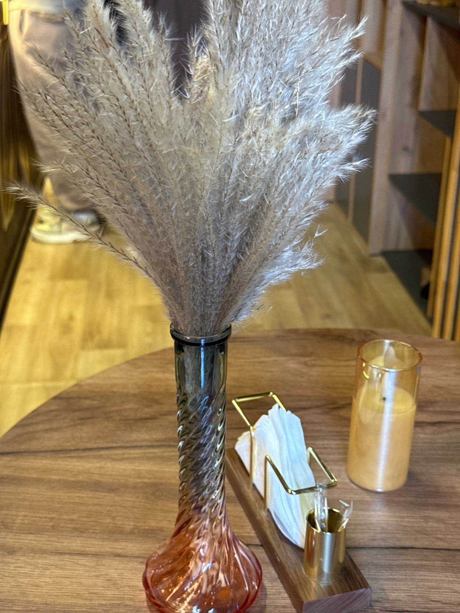An interior scene captures a close-up view of a table setting. In the foreground, a glass vase with a swirling, translucent design, transitioning from a dark top to a reddish-orange base, holds a large, feathery plume of dried grass or reeds. To the right of the vase, a golden metal napkin holder with white napkins is positioned on a wooden base, accompanied by a small gold cup containing what appear to be toothpicks. Further to the right, a cylindrical glass holds a yellowish liquid, possibly a candle or drink. The table surface is made of polished wood with a visible grain. In the blurred background, a person is partially visible wearing light-colored sneakers and dark pants, with a light-colored garment, possibly a handbag, draped over their shoulder. The environment appears to be a cafe or restaurant, with wooden shelves and vertical wooden elements visible in the background. The image is taken in Lyubertsy, Russia.
