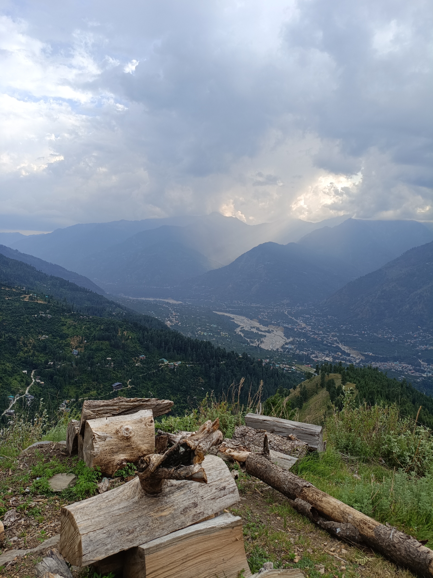 An elevated, overcast view looks out over the valley of Damadi, India, with a village nestled in the distant, hazy mountains. The foreground is dominated by a collection of weathered, rough-hewn logs stacked on a grassy slope. Beyond the logs, the verdant, terraced hillsides are dotted with small homes, and further in the distance, a winding river cuts through the valley floor. The sky is a dramatic mix of grey clouds with shafts of sunlight breaking through, illuminating parts of the mountain range.