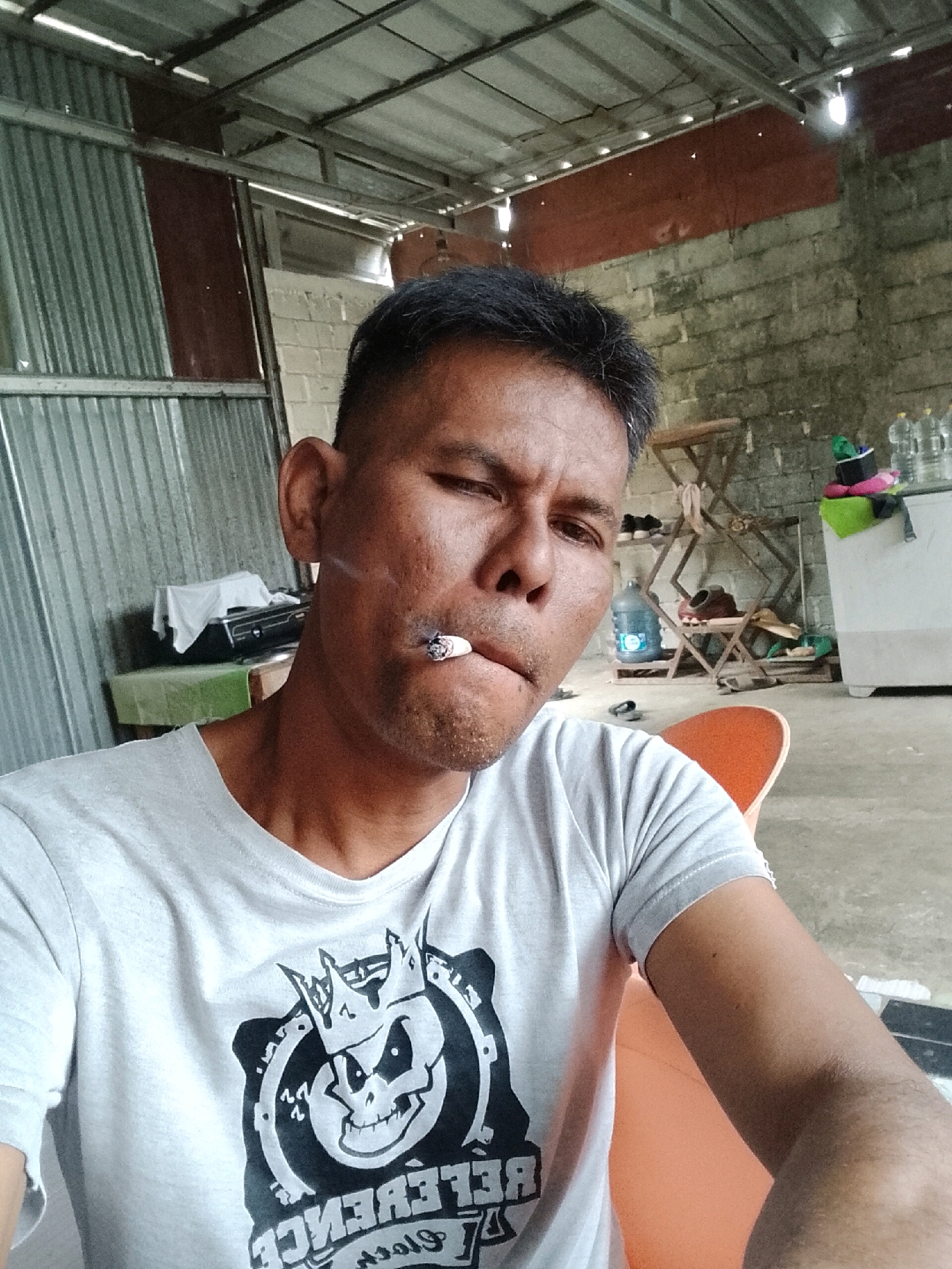 An adult male with short, dark hair and an olive complexion is pictured in a close-up, selfie-style shot. He is wearing a light grey t-shirt with a black graphic design on the front. A lit cigarette is held between his lips, with smoke visible. The man's face is turned slightly to the left, and his eyes are partially closed.
The background suggests an open-air, possibly unfinished, structure. A corrugated metal roof is visible overhead, supported by metal beams. The walls appear to be constructed of grey cinder blocks. To the left, a section of grey corrugated metal siding is present. In the mid-ground, to the left of the man, is a green-covered table with a white cloth draped over an object on it. Behind the table, a black electronic device is visible.
Further into the background, on the right side of the image, is a white cabinet or counter. Various items are placed on and around it, including several plastic water bottles, a green object, and other unidentifiable items. A metal folding rack with shelves is also visible in the background, holding a water jug, a black pot, and other assorted objects. The floor appears to be concrete.
The scene is consistent with a residential or workshop area in a tropical climate, potentially in a rural or semi-urban setting like Padang Dalom, Indonesia. The overall impression is informal and functional.